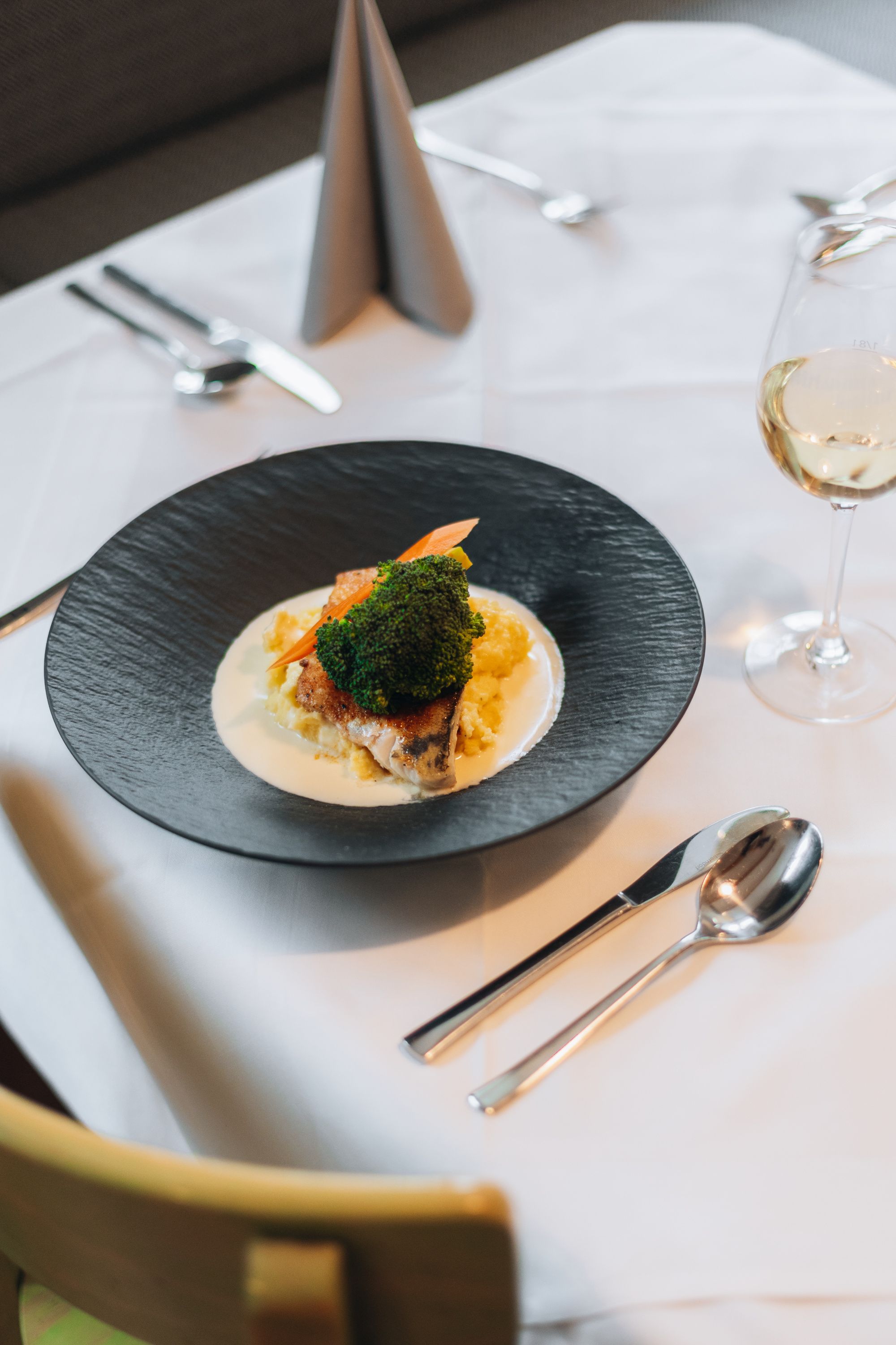 Elegantně prezentovaný pokrm na černém talíři se zeleninou a omáčkou, doplněný sklenkou bílého vína na prostřeném stole.