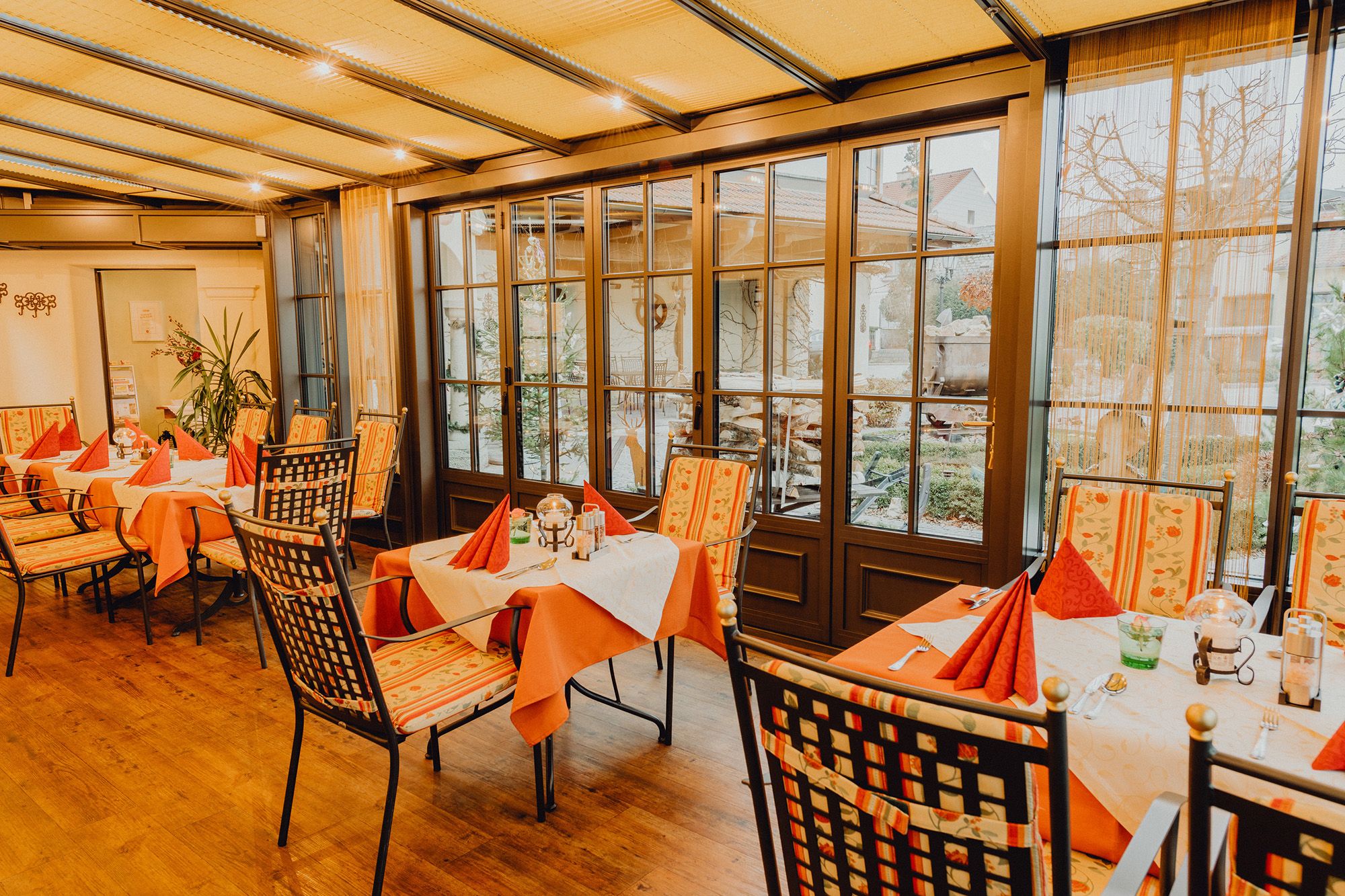 Útulná zimní zahrada se stoly a židlemi, obklopená velkými okny s výhledem ven.