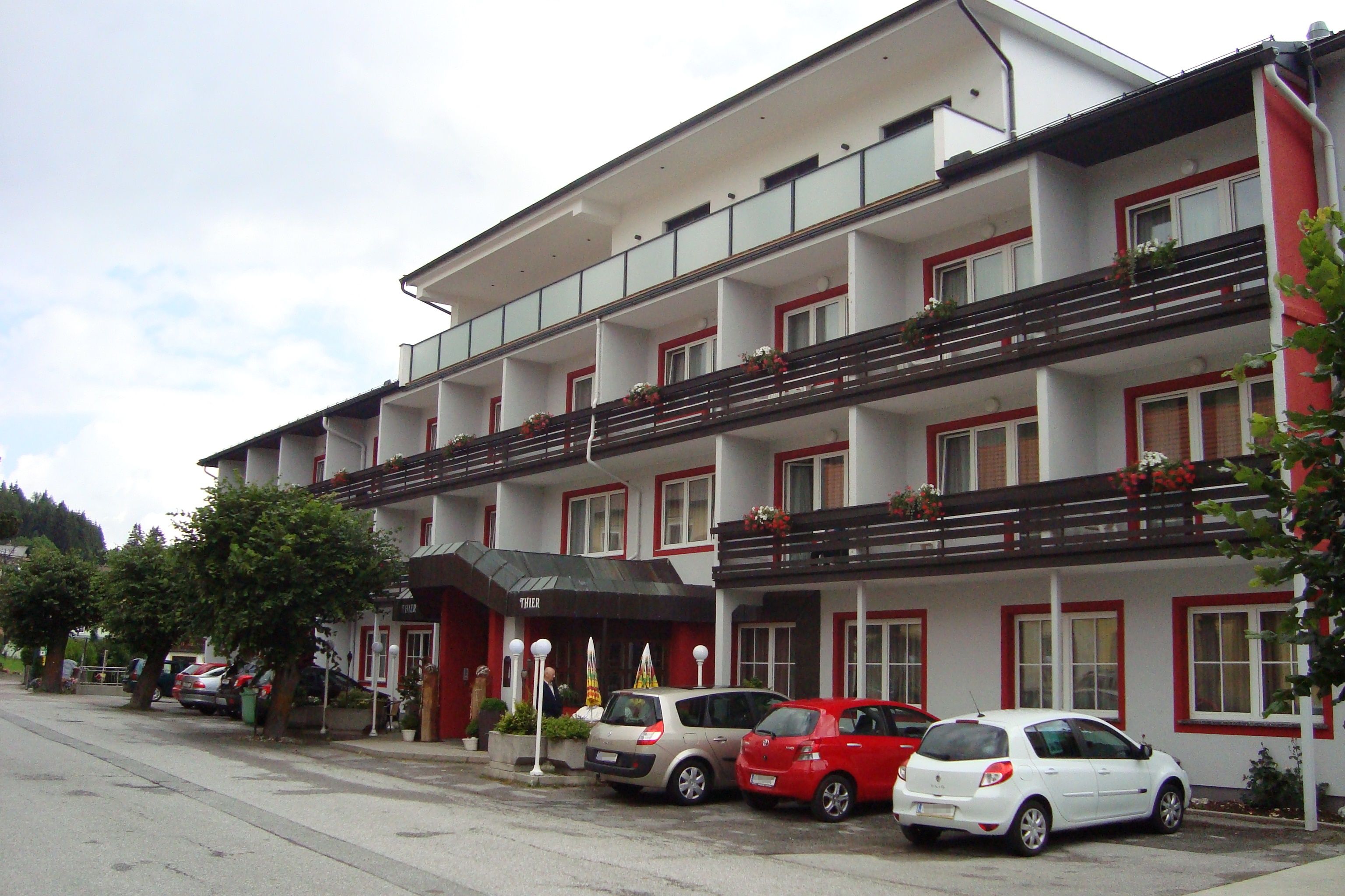 Pohled na exteriér hotelu Thier se zaparkovanými auty a balkony s květinami.