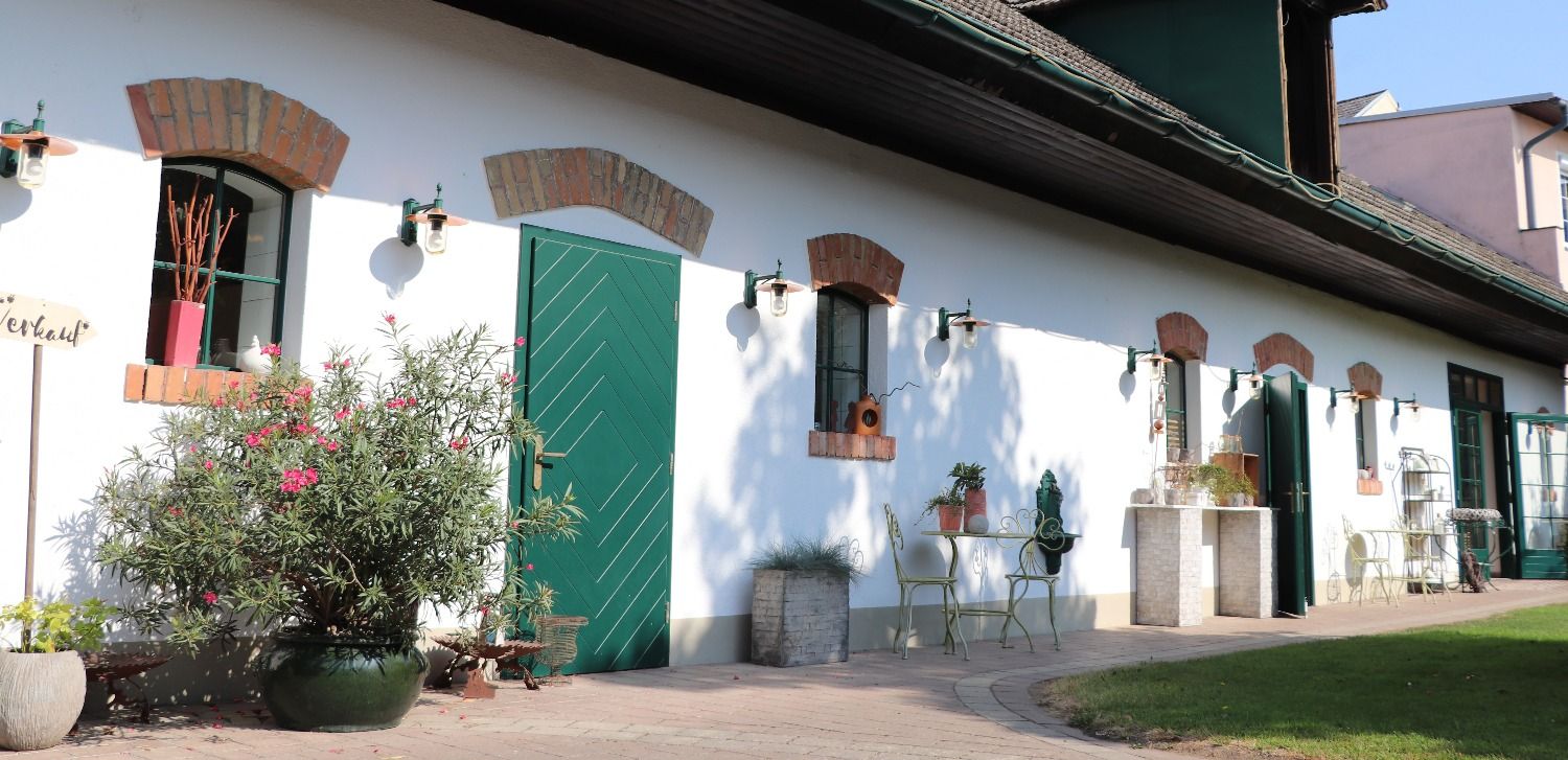 Bílá budova se zelenými dveřmi a okny, obklopená rostlinami a zahradním nábytkem.