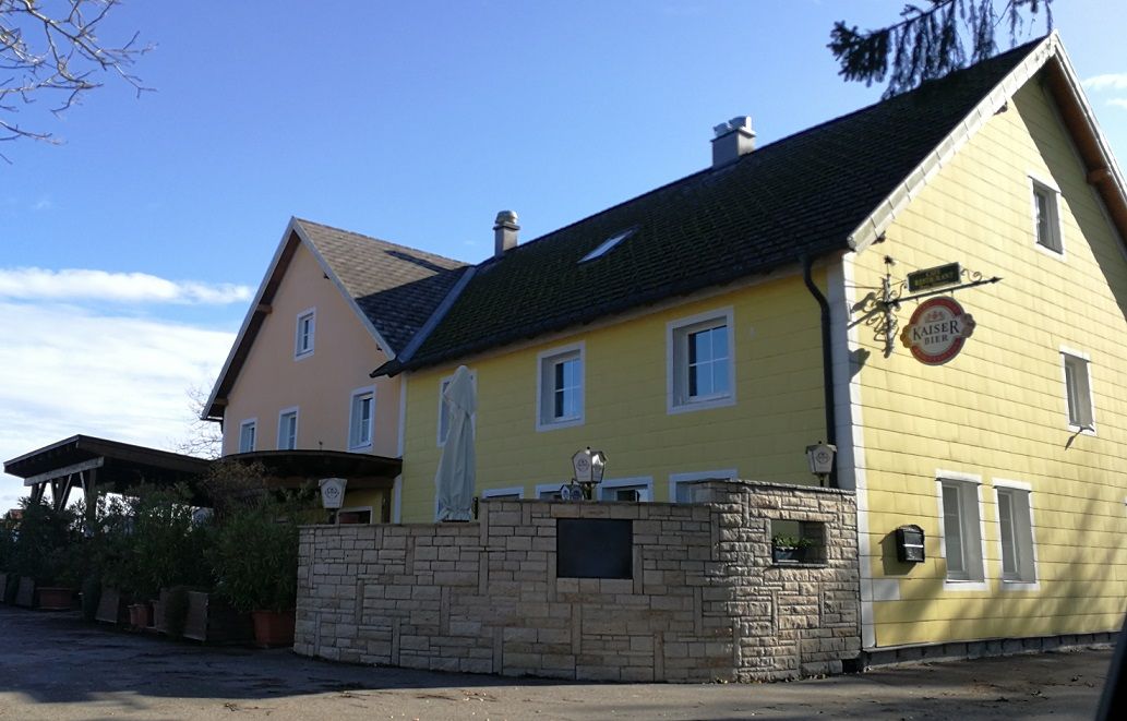 Žlutý venkovský hostinec s nápisem Kaiser Bier a slunečníkem v popředí.