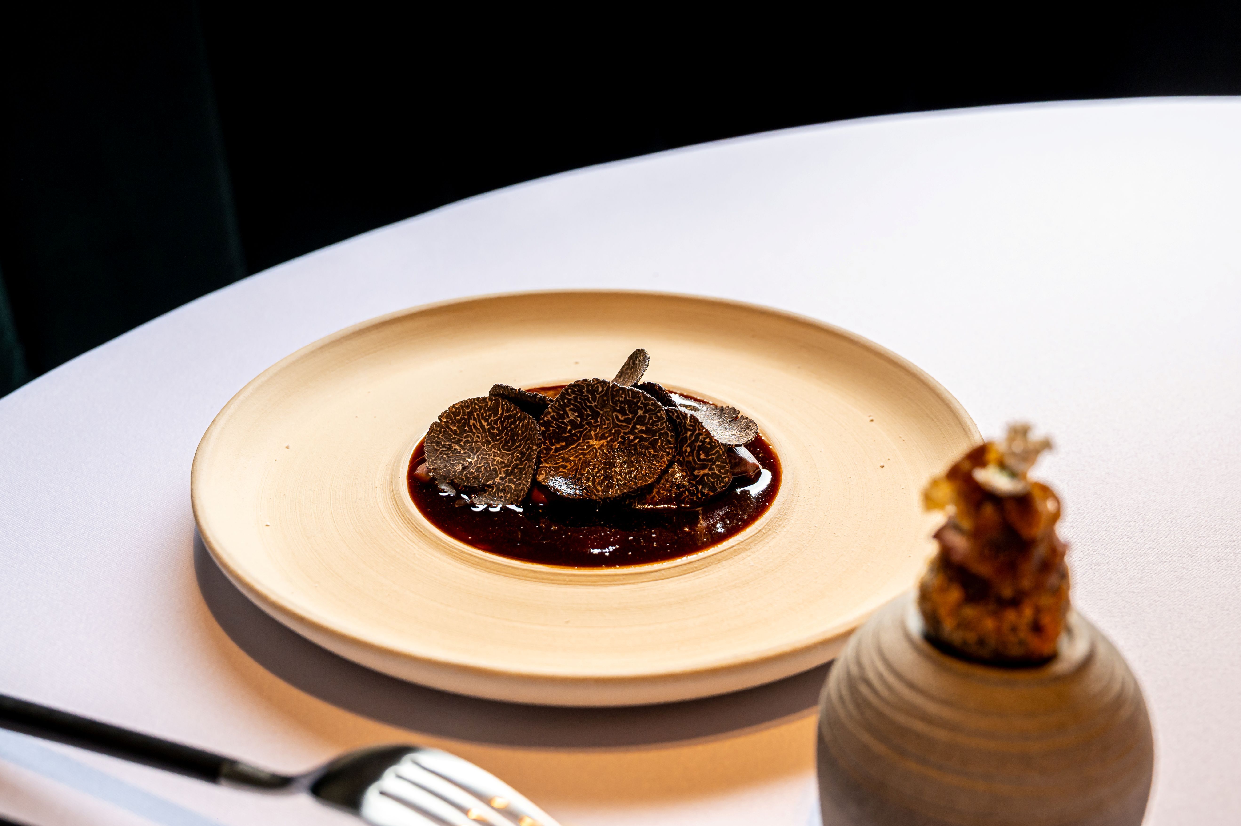 Gurmánský pokrm s plátky lanýžů na talíři, vedle něhož je umístěn dekorativní prvek.