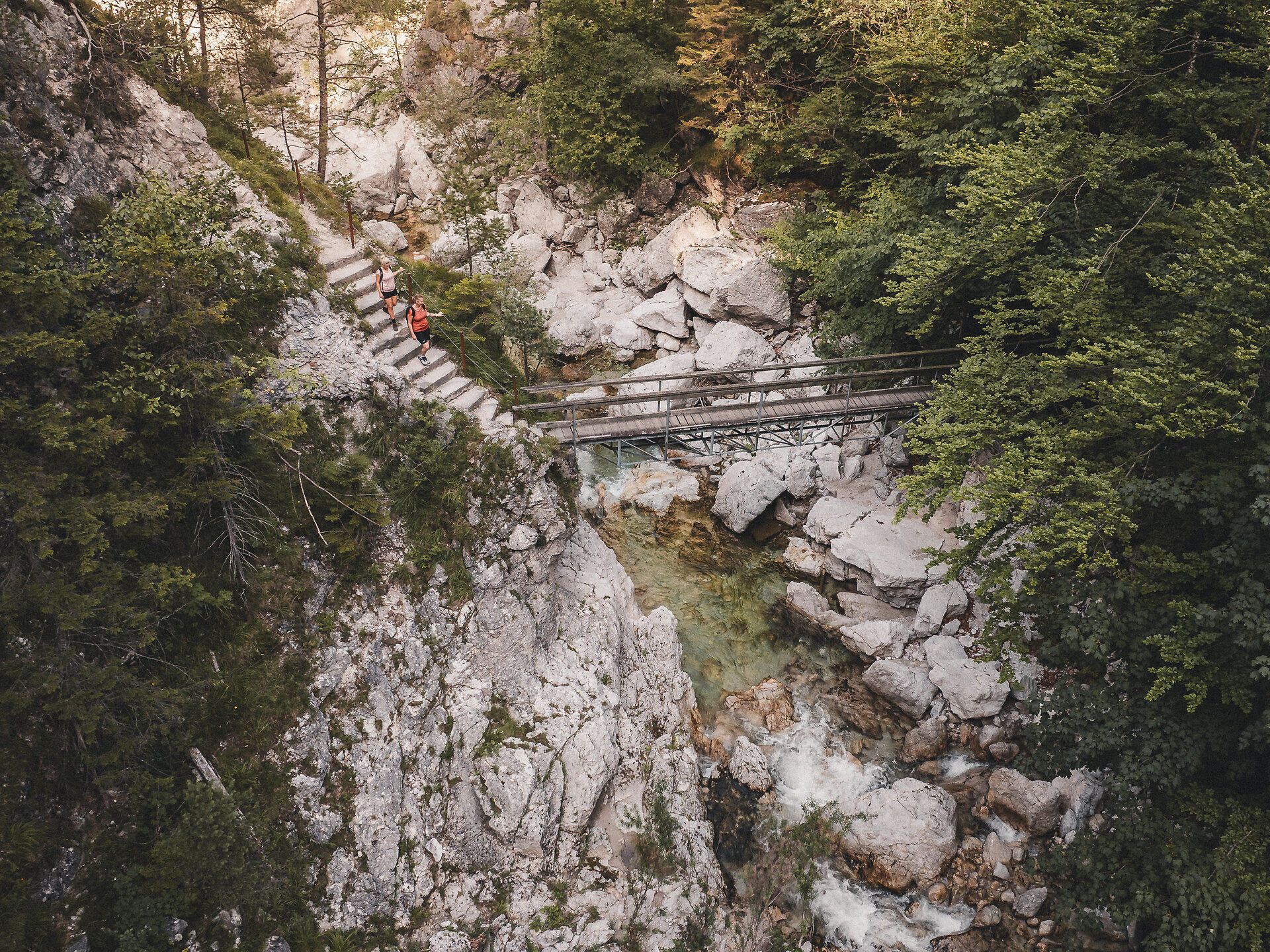 Zwei Wanderinnen überqueren eine Holzbrücke über einen felsigen Bachlauf in einer bewaldeten Schlucht