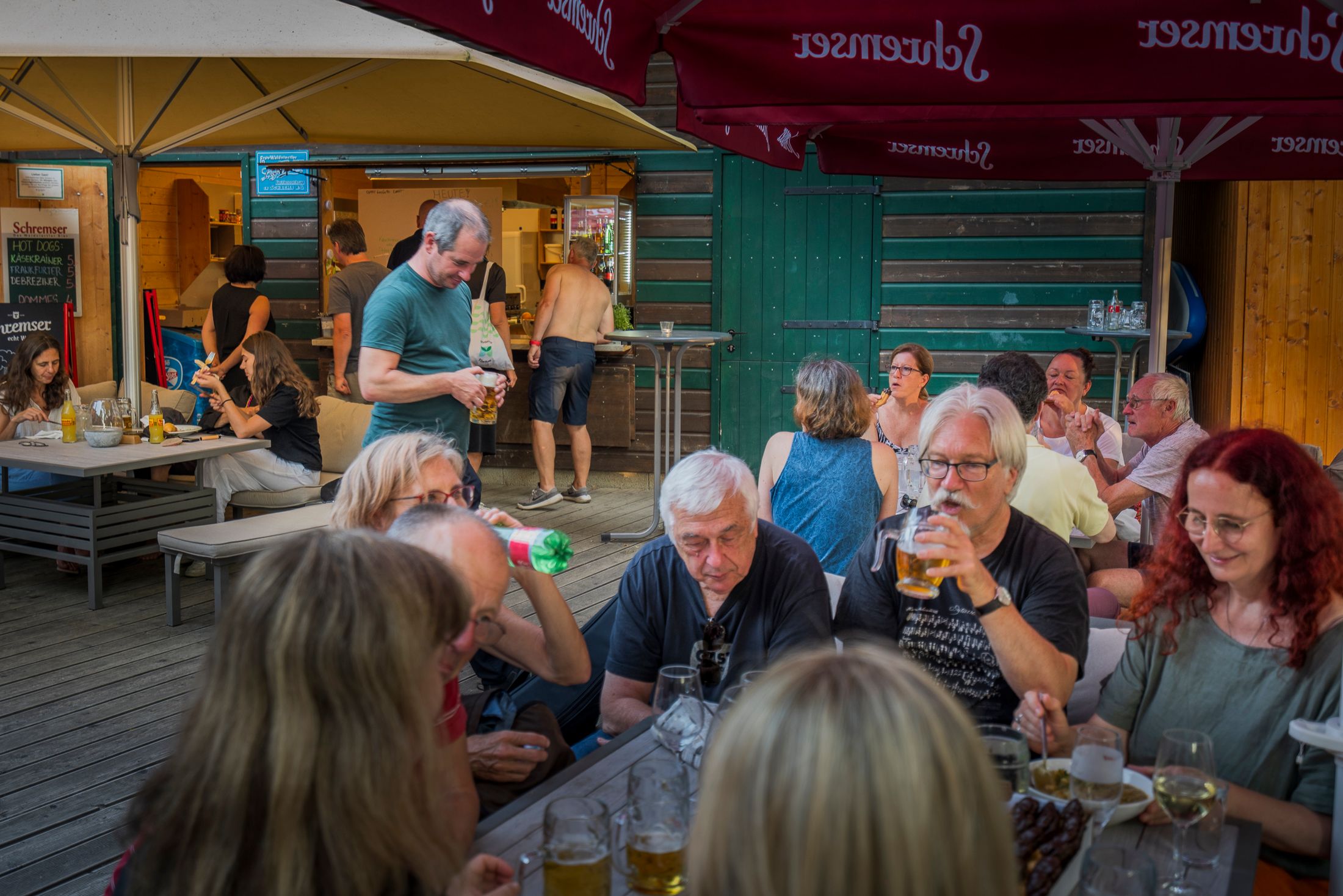 Lidé sedí u stolů v pivní zahrádce a vychutnávají si nápoje a občerstvení.