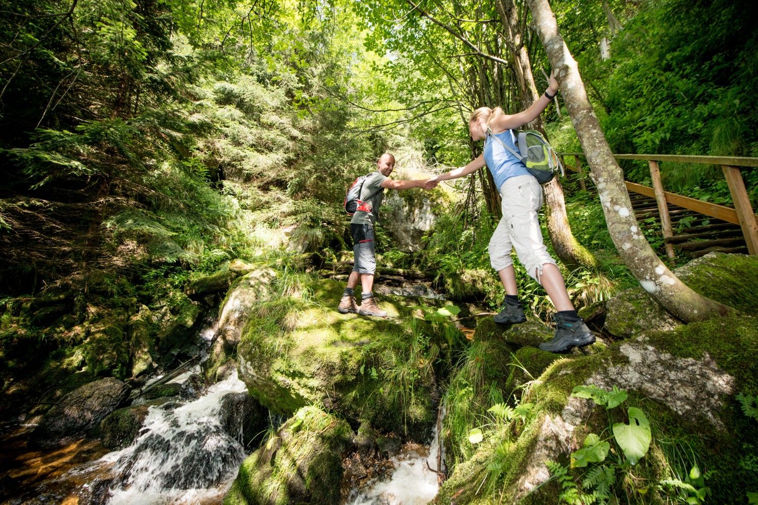 Dva turisté si pomáhají přejít potok v zeleném lese.