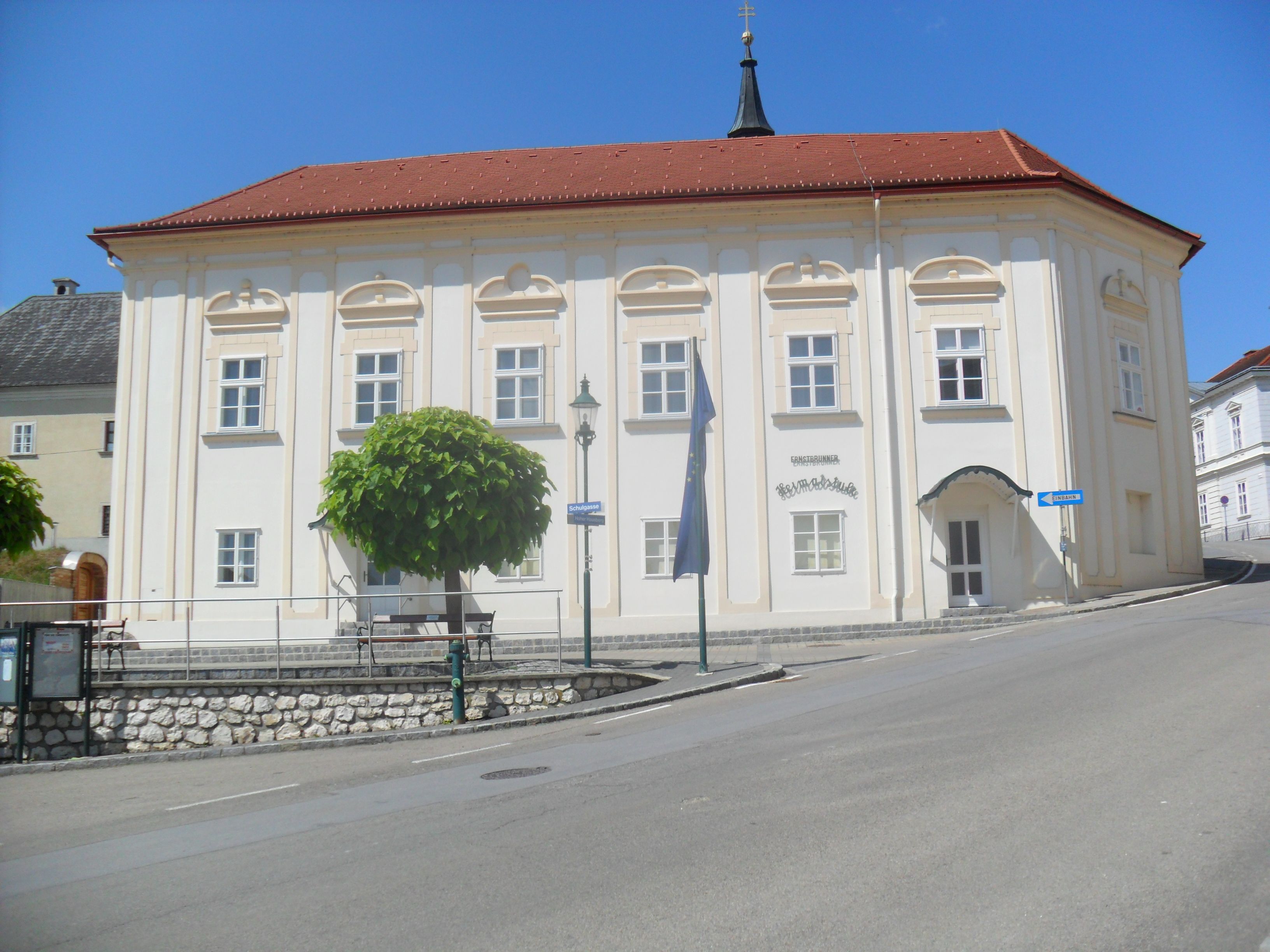 Historická budova s červenou střechou a bílými zdmi, obklopená stromy a silnicí.