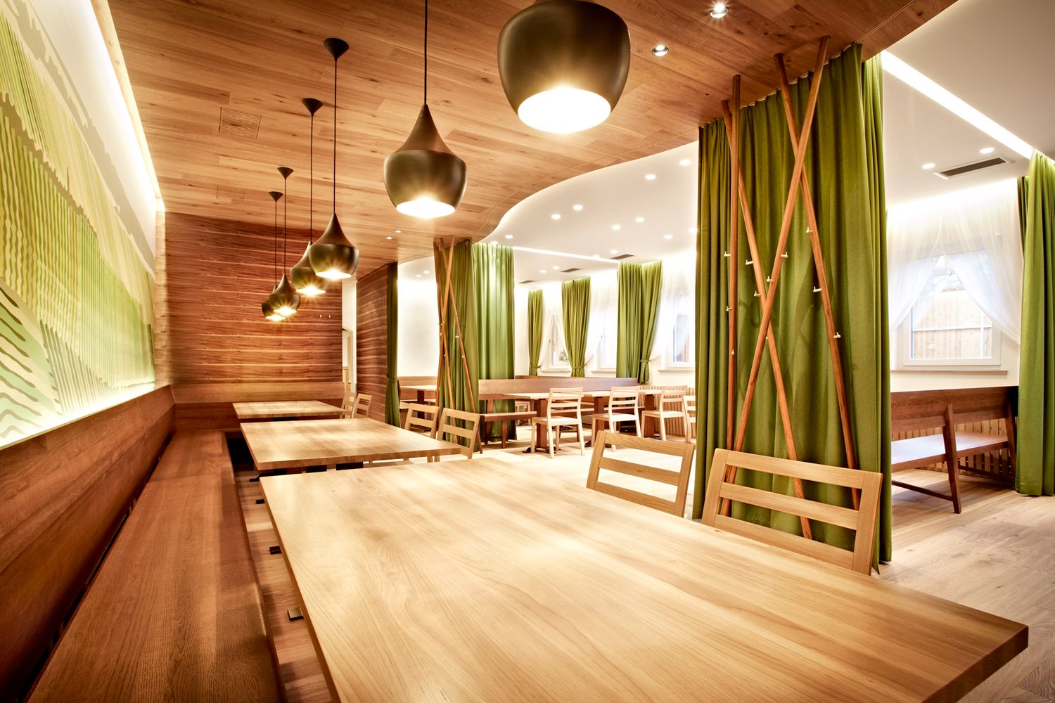 Moderní restaurace s dřevěnými stoly a zelenými závěsy.