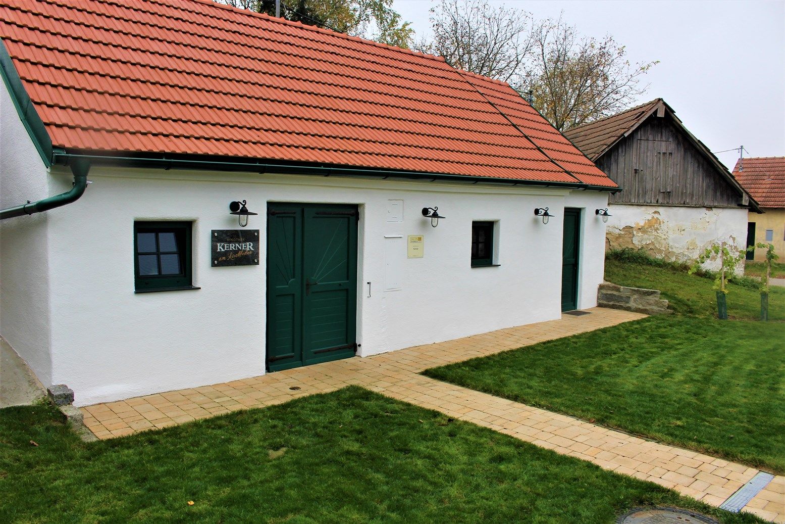 Bílá budova s červenou střechou a zelenými dveřmi, obklopená trávníkem.