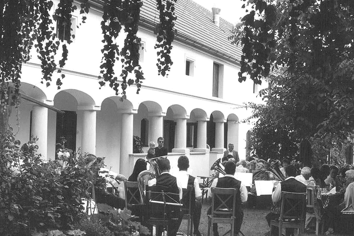 Skupina lidí sedí venku před budovou se sloupy a hraje dechovka.