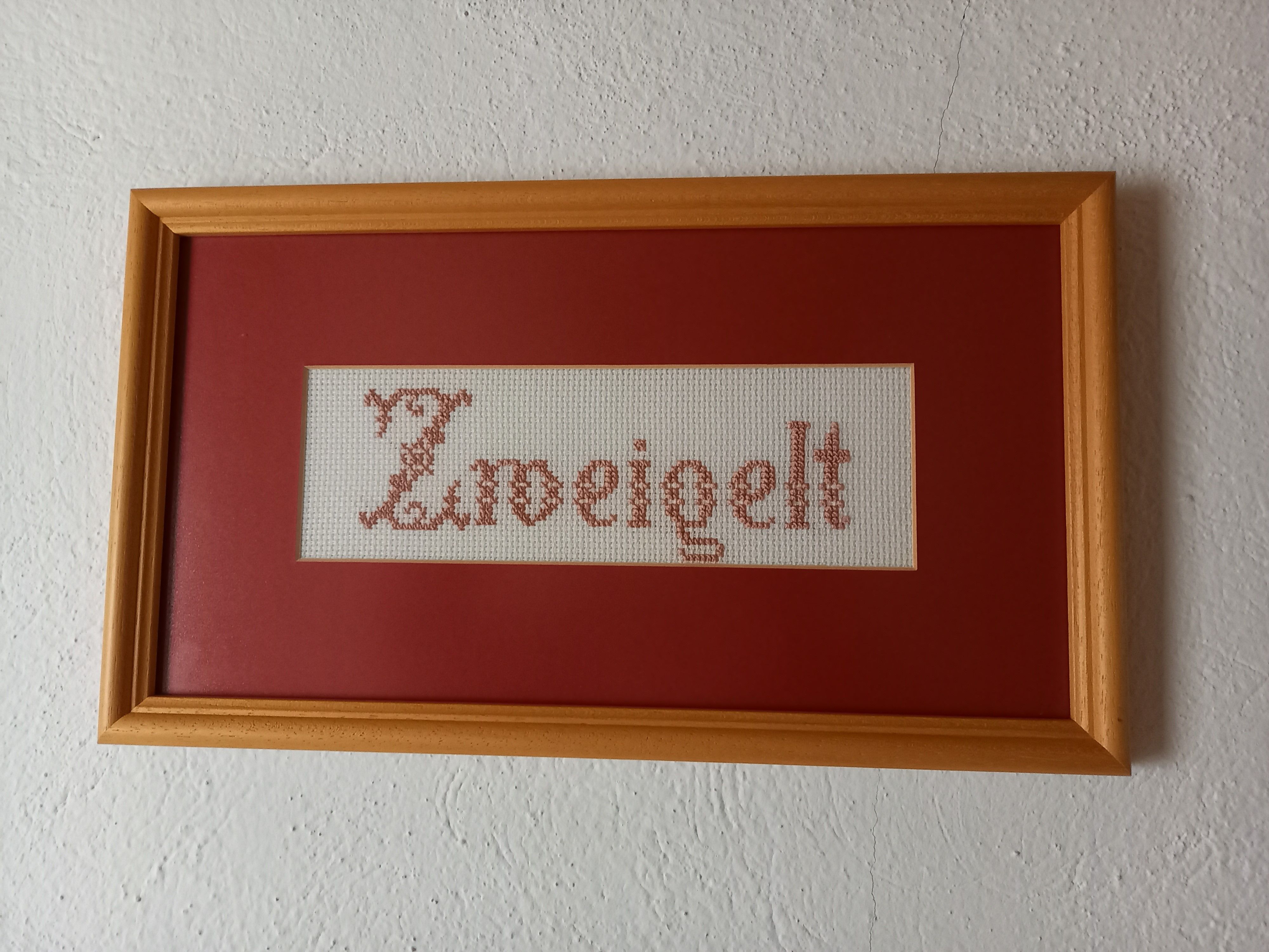 Vyšívaný obrázek s nápisem "Zweigelt" v dřevěném rámečku.