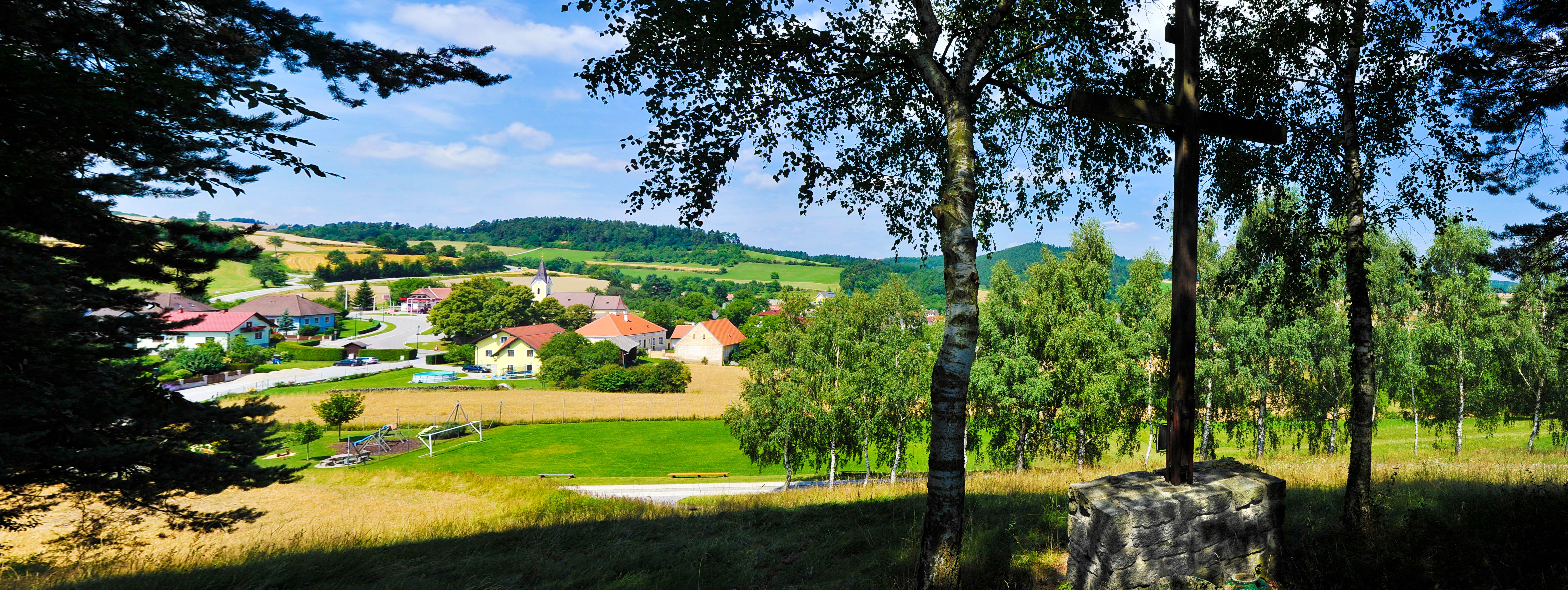 Pohled na venkovskou obec s kostelem a domy, obklopenou poli a stromy.