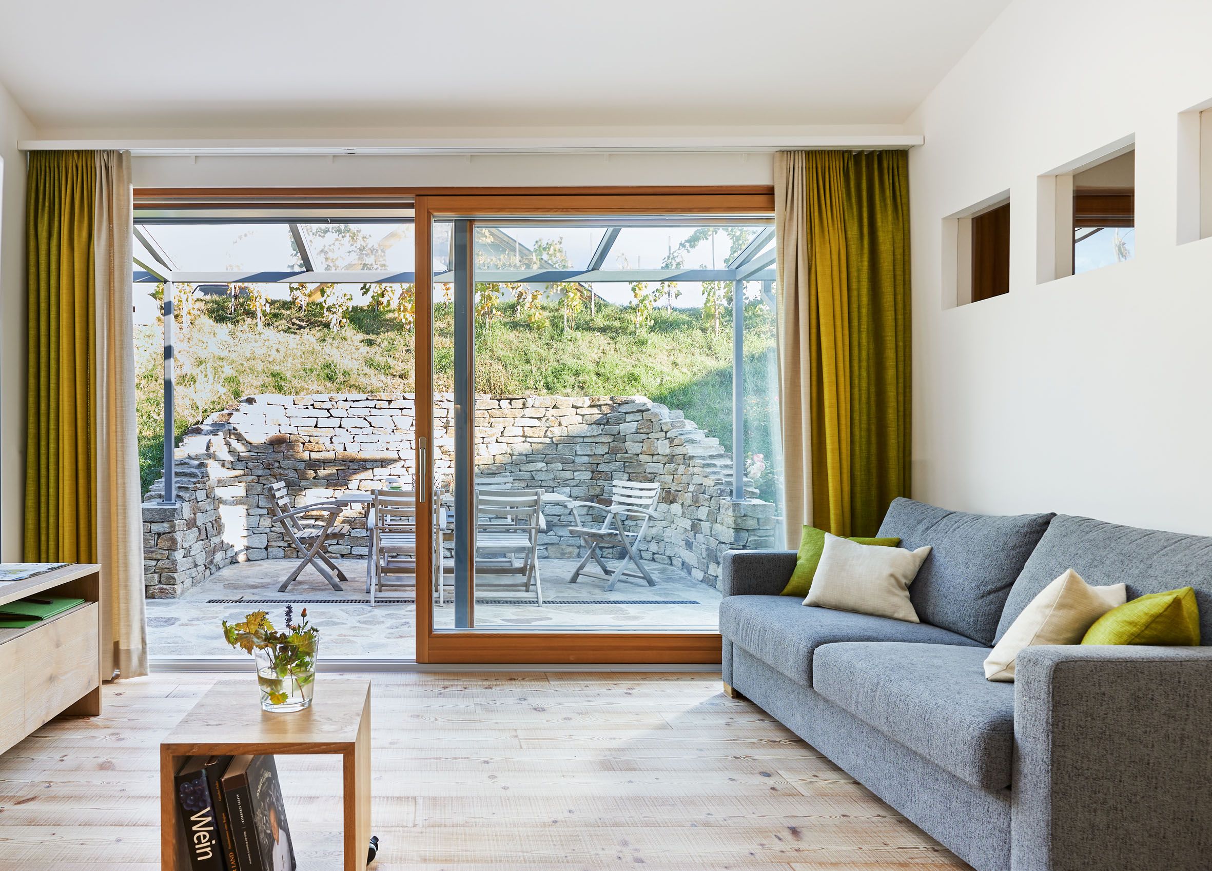 Moderní obývací pokoj s šedou pohovkou, dřevěnou podlahou a velkými okny s výhledem na terasu s kamennou zdí a zahradním nábytkem.