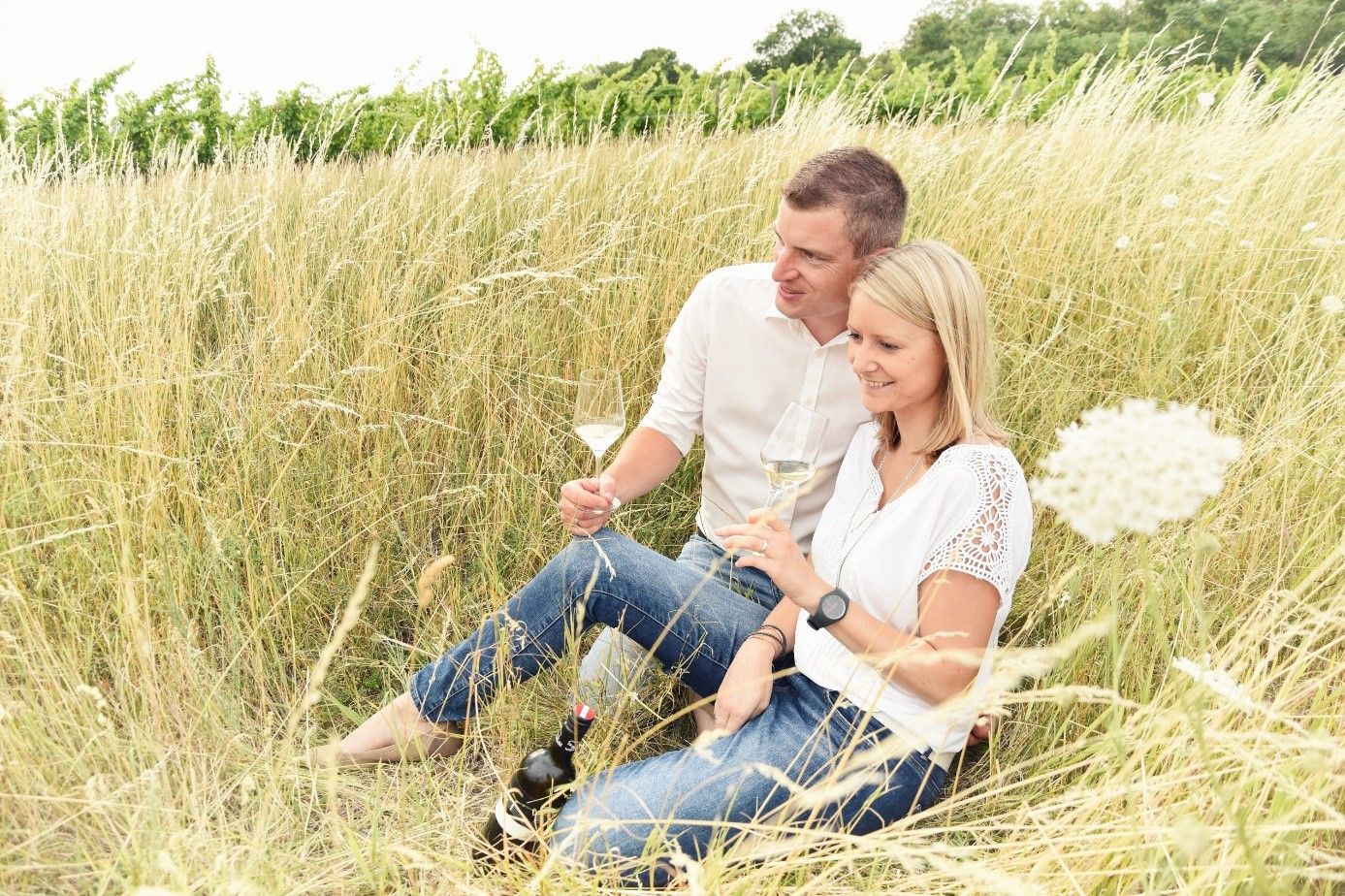 Pár sedí ve vysoké trávě a popíjí víno.