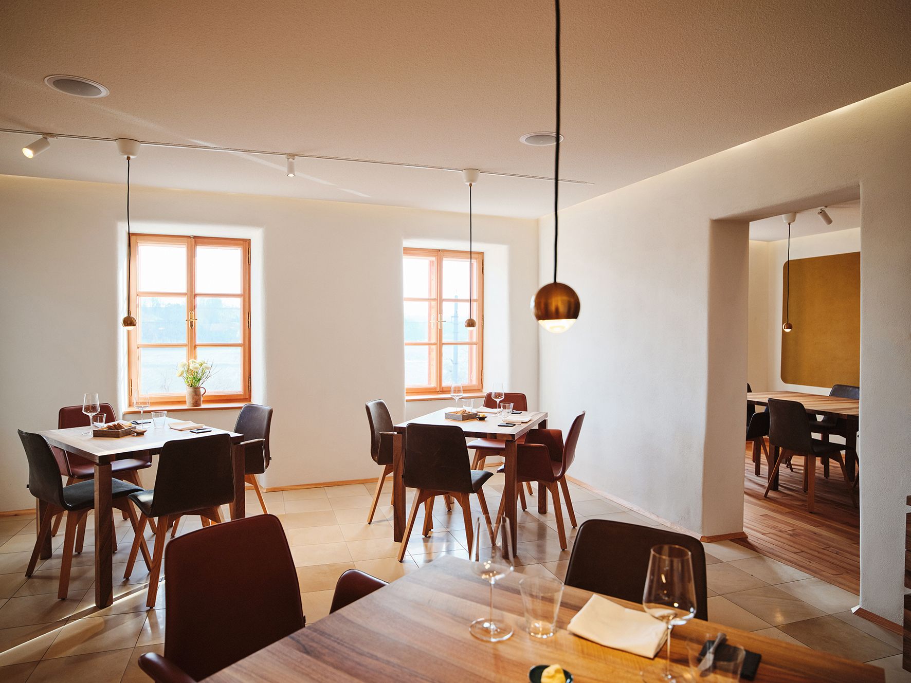 Moderní jídelna s dřevěnými stoly, sklenicemi na víno a velkými okny v minimalistickém designu.