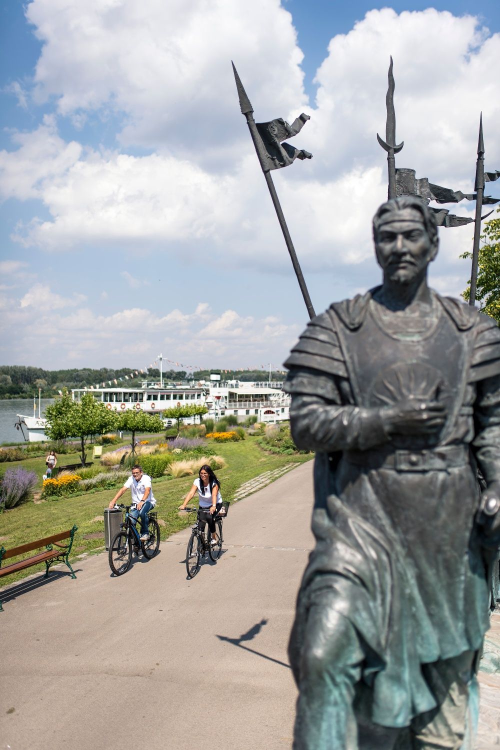 Dva cyklisté jedou po stezce podél Dunaje v Tullnu. V popředí je socha a v pozadí je vidět říční výletní loď.