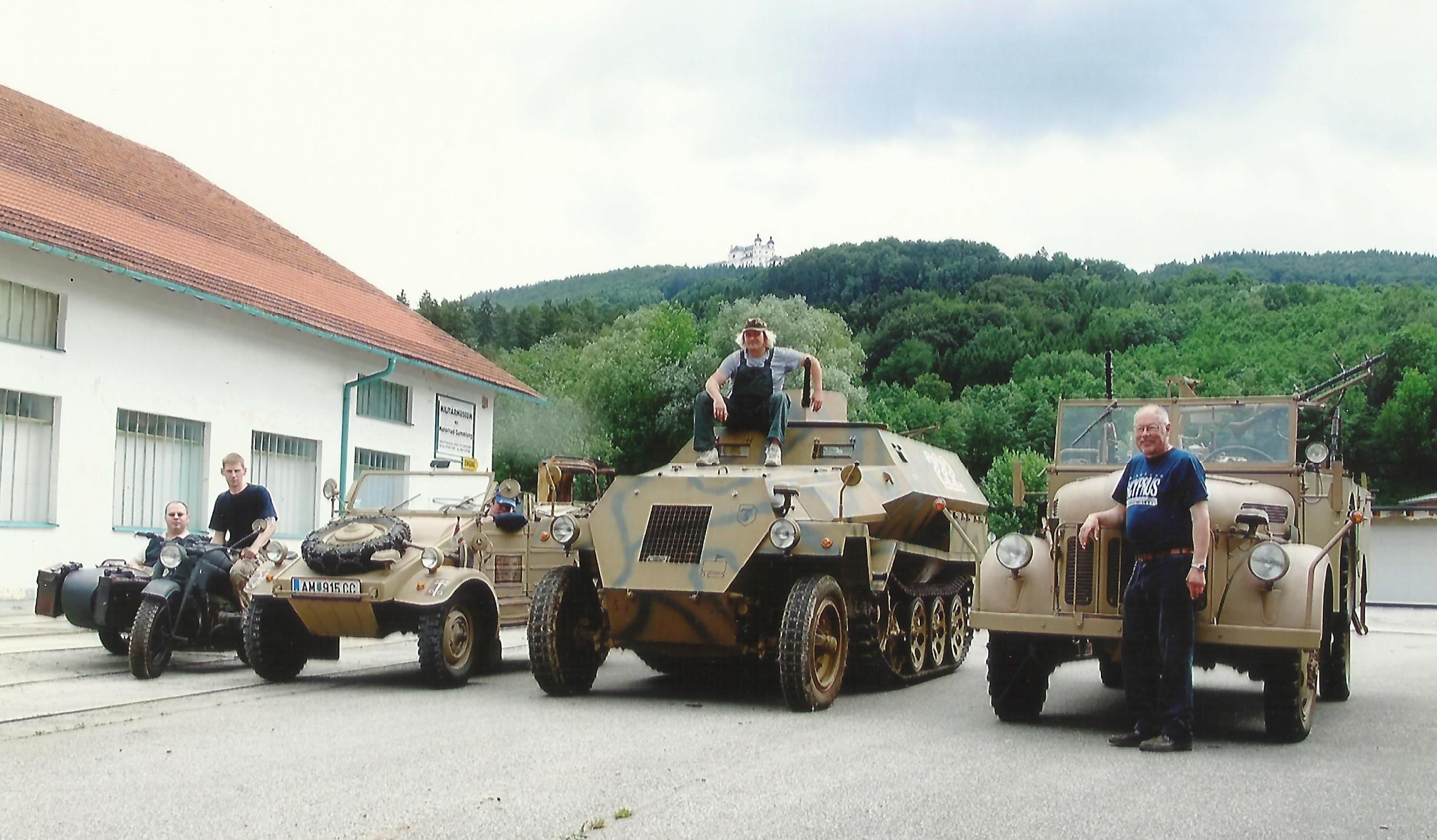 Vojenská vozidla před budovou s kopcem v pozadí.