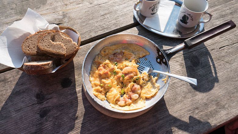 Pánev s míchanými vejci a slaninou na dřevěném stole, vedle košík na chleba a dva hrnky.