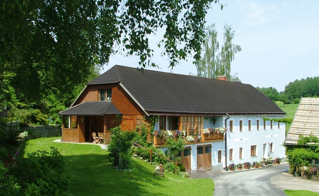 Tradiční statek s květinami na balkoně, obklopený zelenou krajinou a stromy.