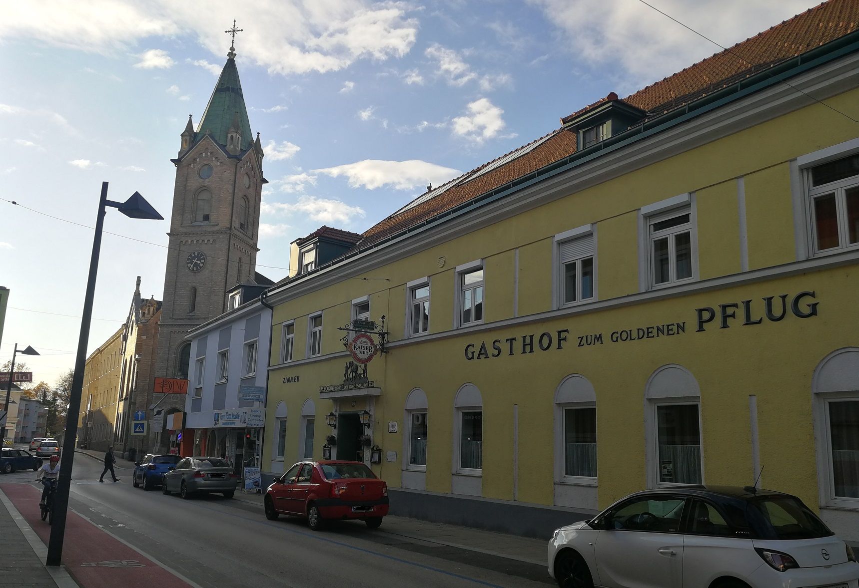 Uliční scéna se žlutou budovou "Gasthof zum goldenen Pflug" a kostelní věží v pozadí.