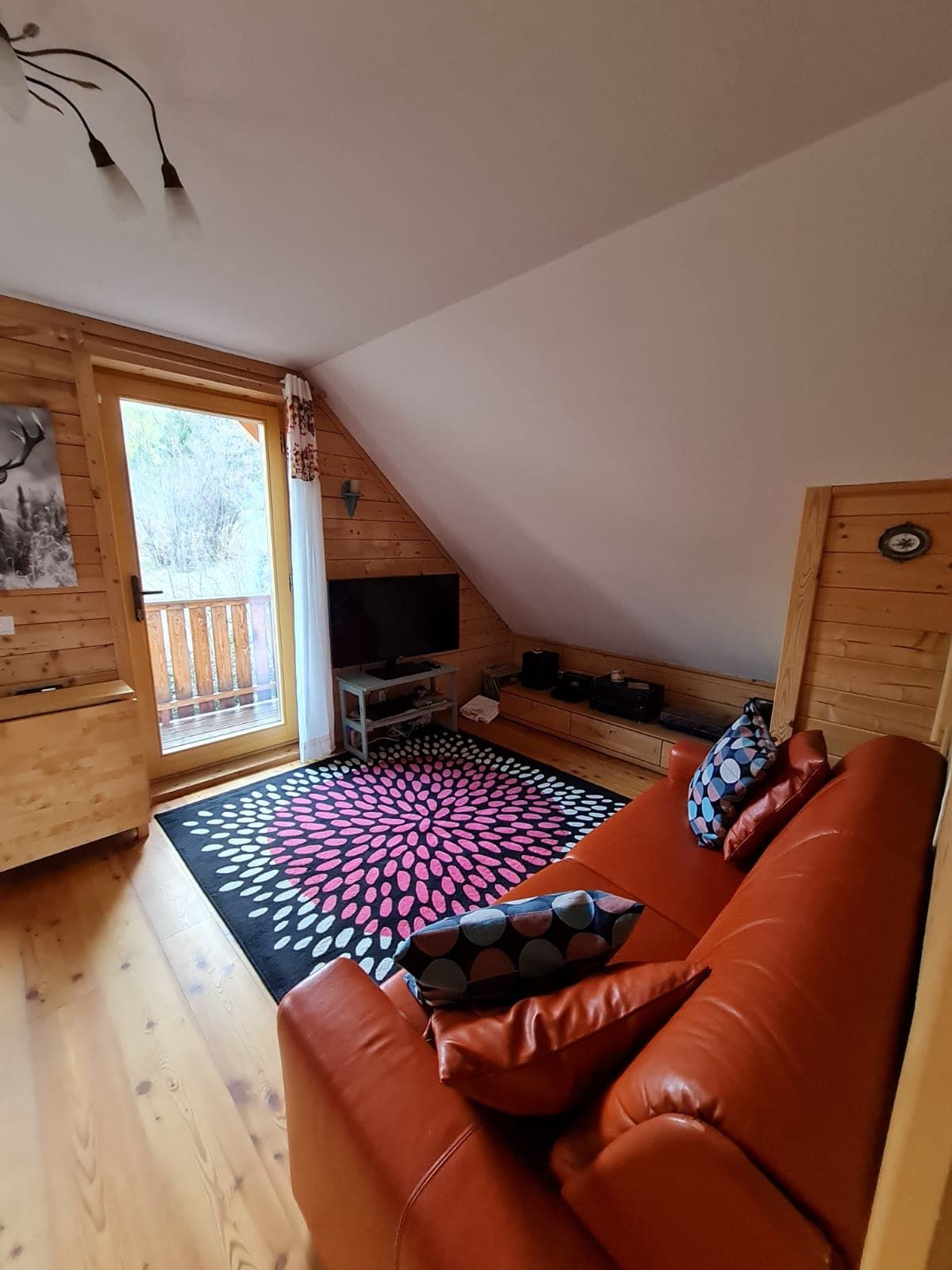 Útulný obývací pokoj s červenou pohovkou, dřevěnými stěnami a barevným kobercem před balkonovým oknem.