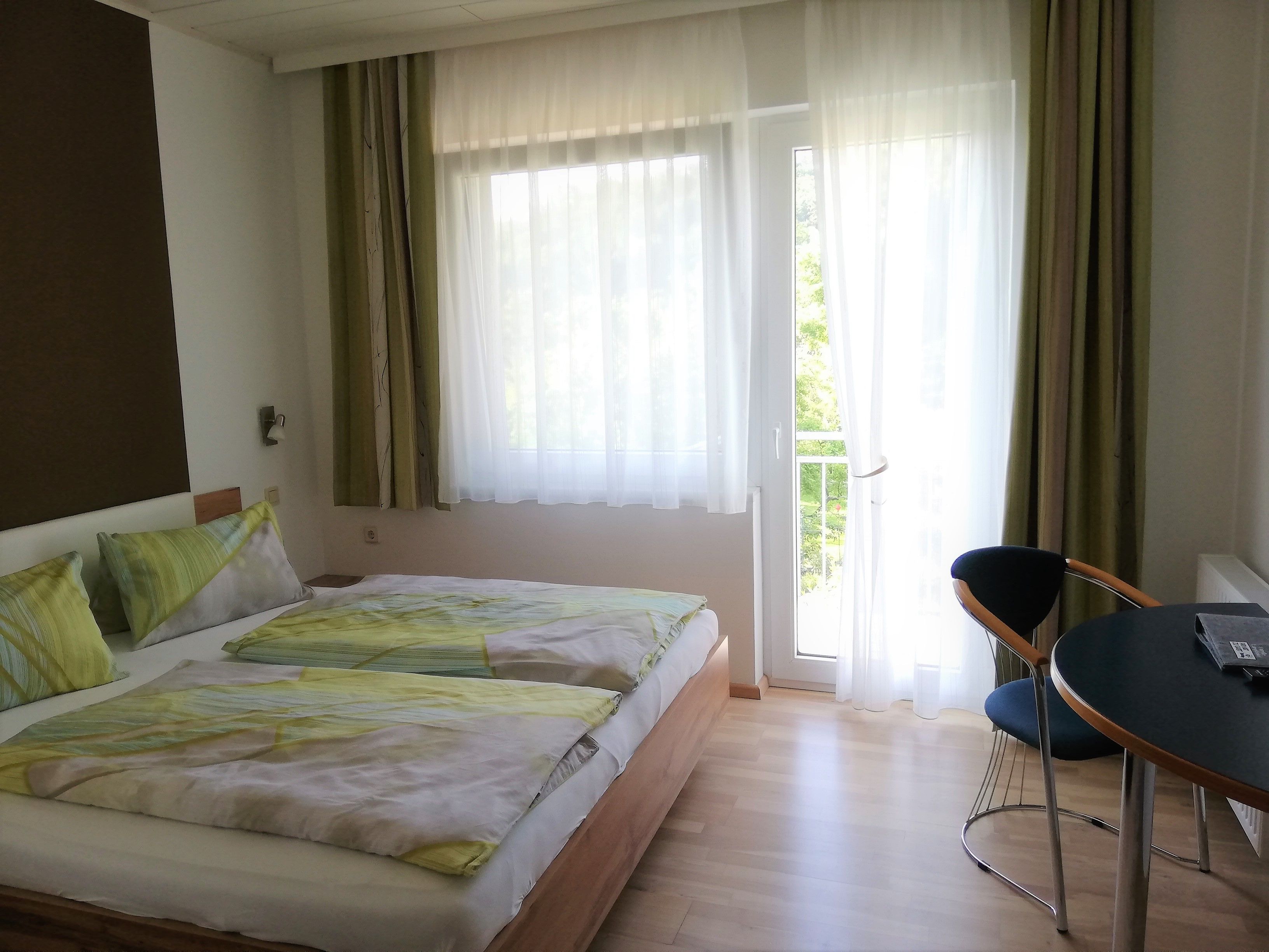 Dvoulůžkový pokoj s postelí, stolem a židlí. Skleněnými dveřmi je vidět na balkon.