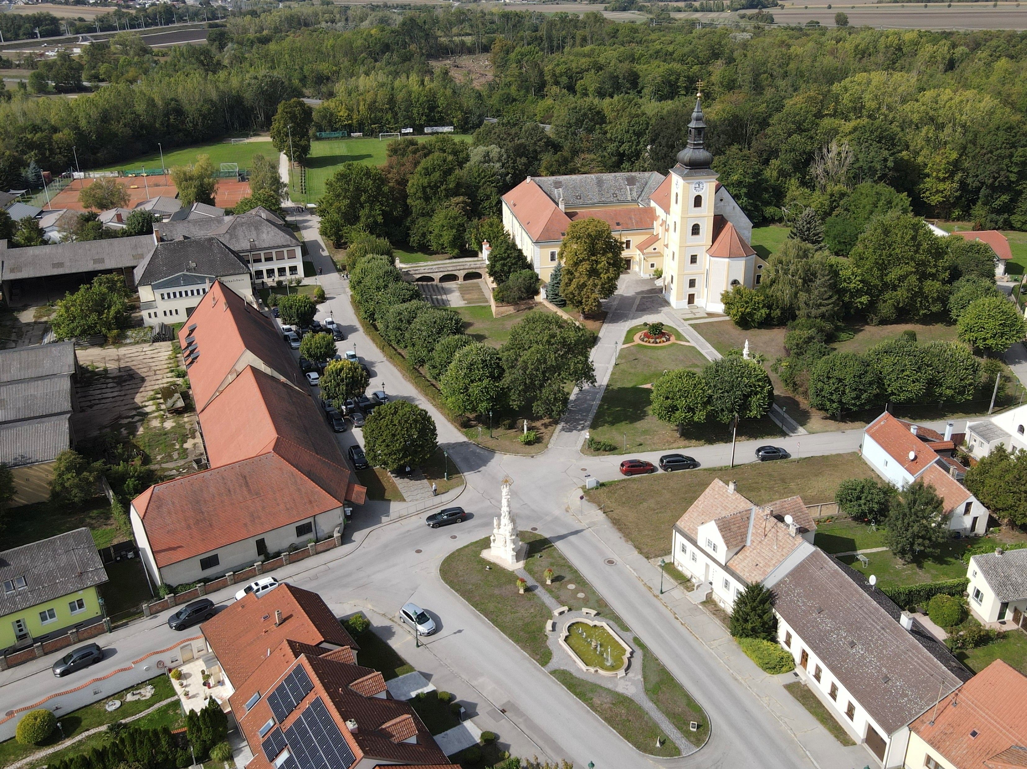 Letecký snímek Weikendorfu s kostelem, návsí, fontánou a okolními domy na okraji lesního porostu.