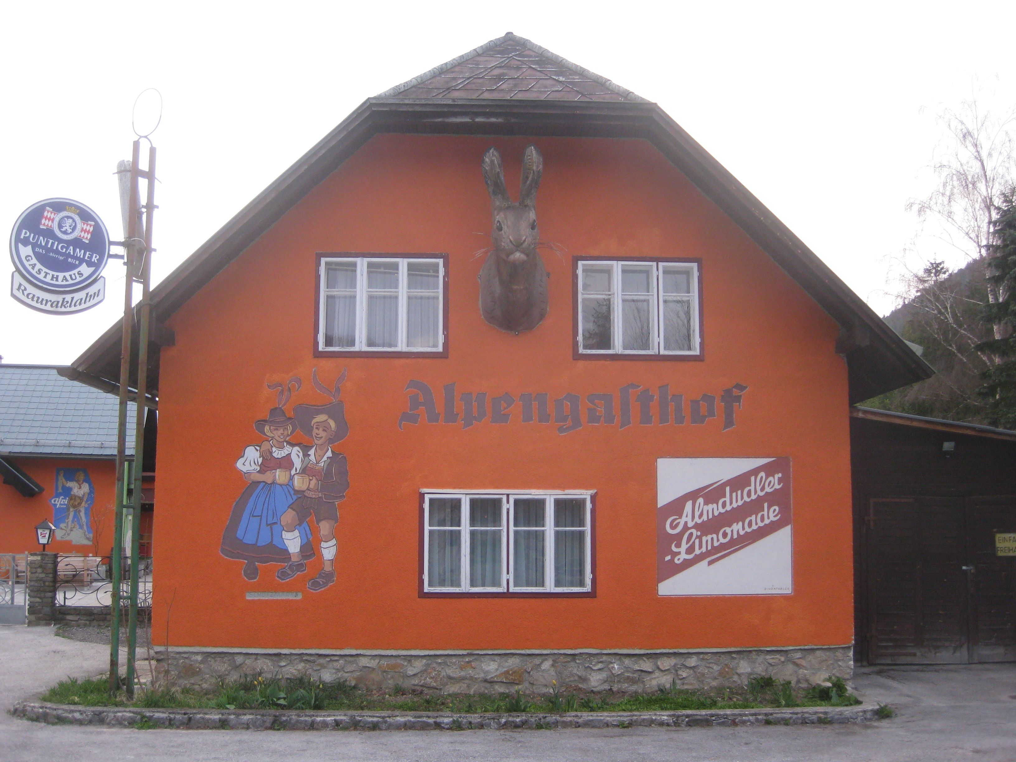 Oranžově natřená budova s nápisem "Alpengasthof", nástěnná malba tančícího páru a reklama Almdudler.