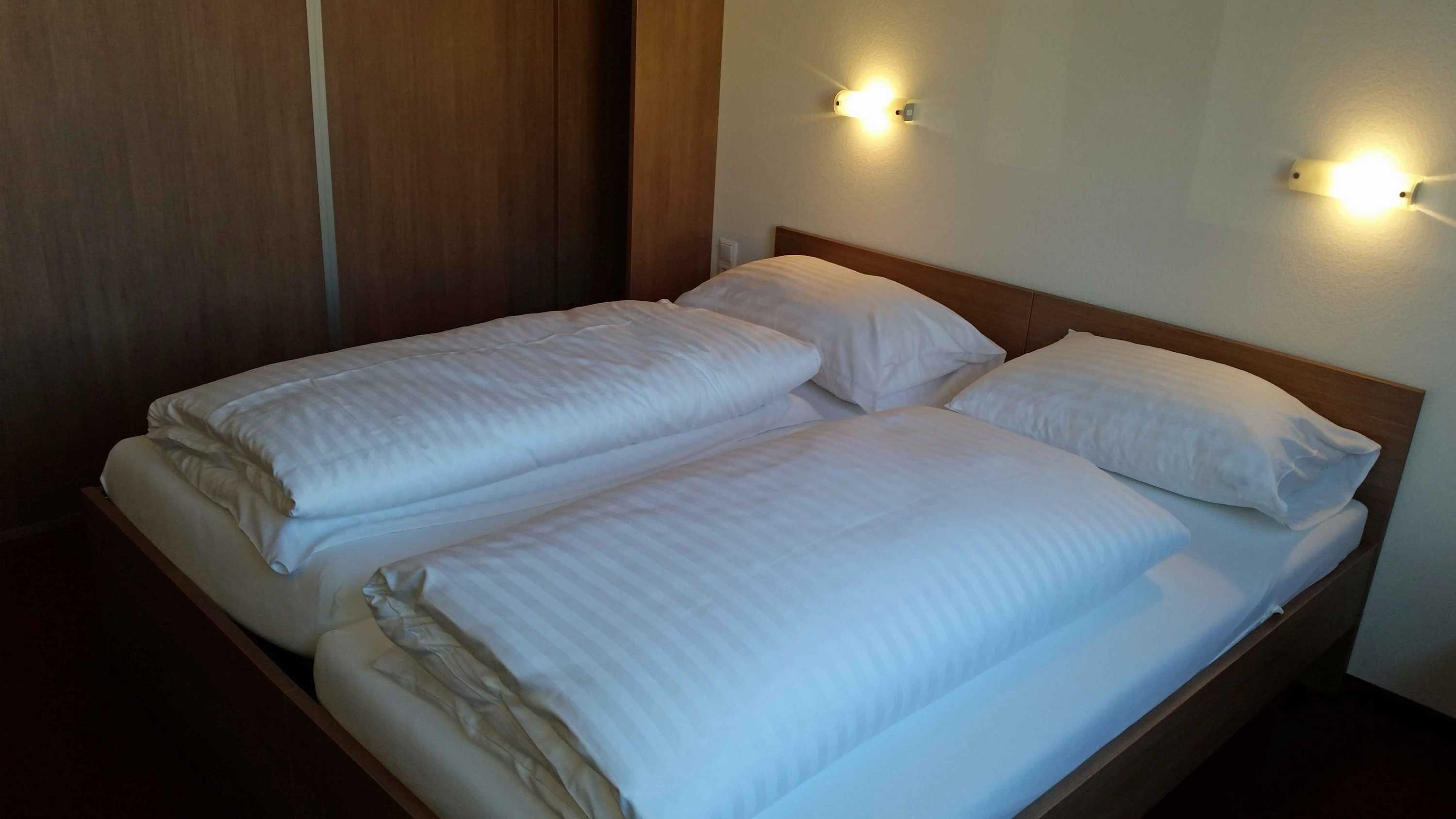 Manželská postel v motelovém pokoji s bílými peřinami a polštáři, osvětlená dvěma nástěnnými lampami.