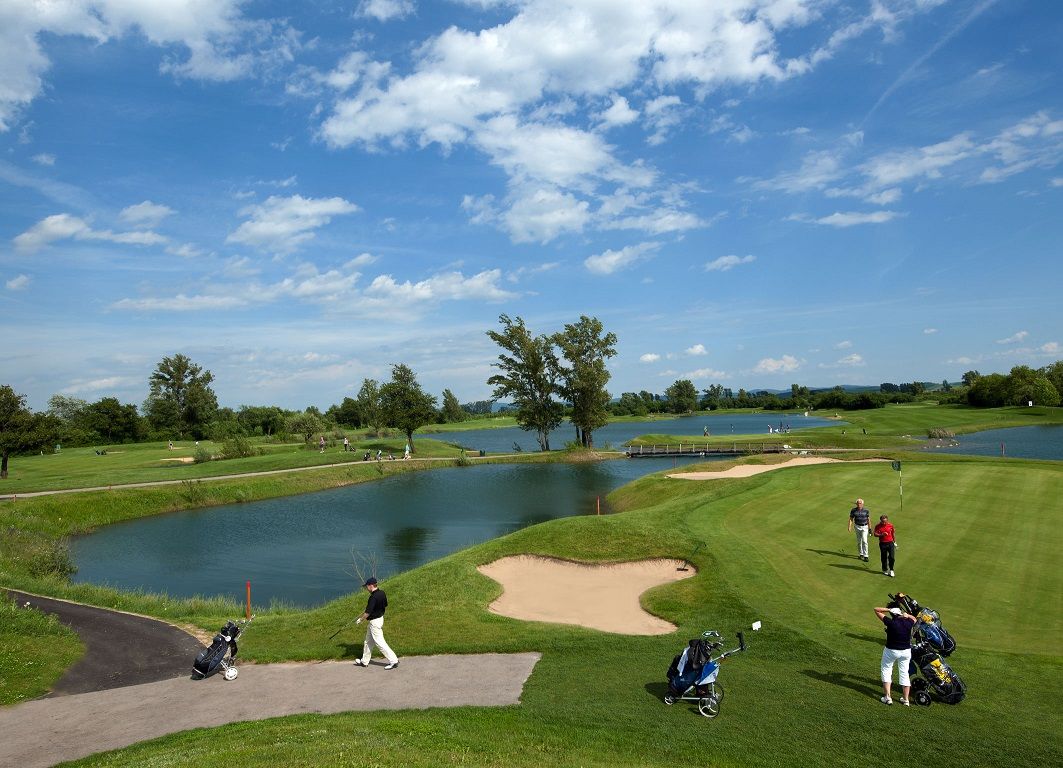 Golfové hřiště s vodními překážkami a hráči pod modrou oblohou.