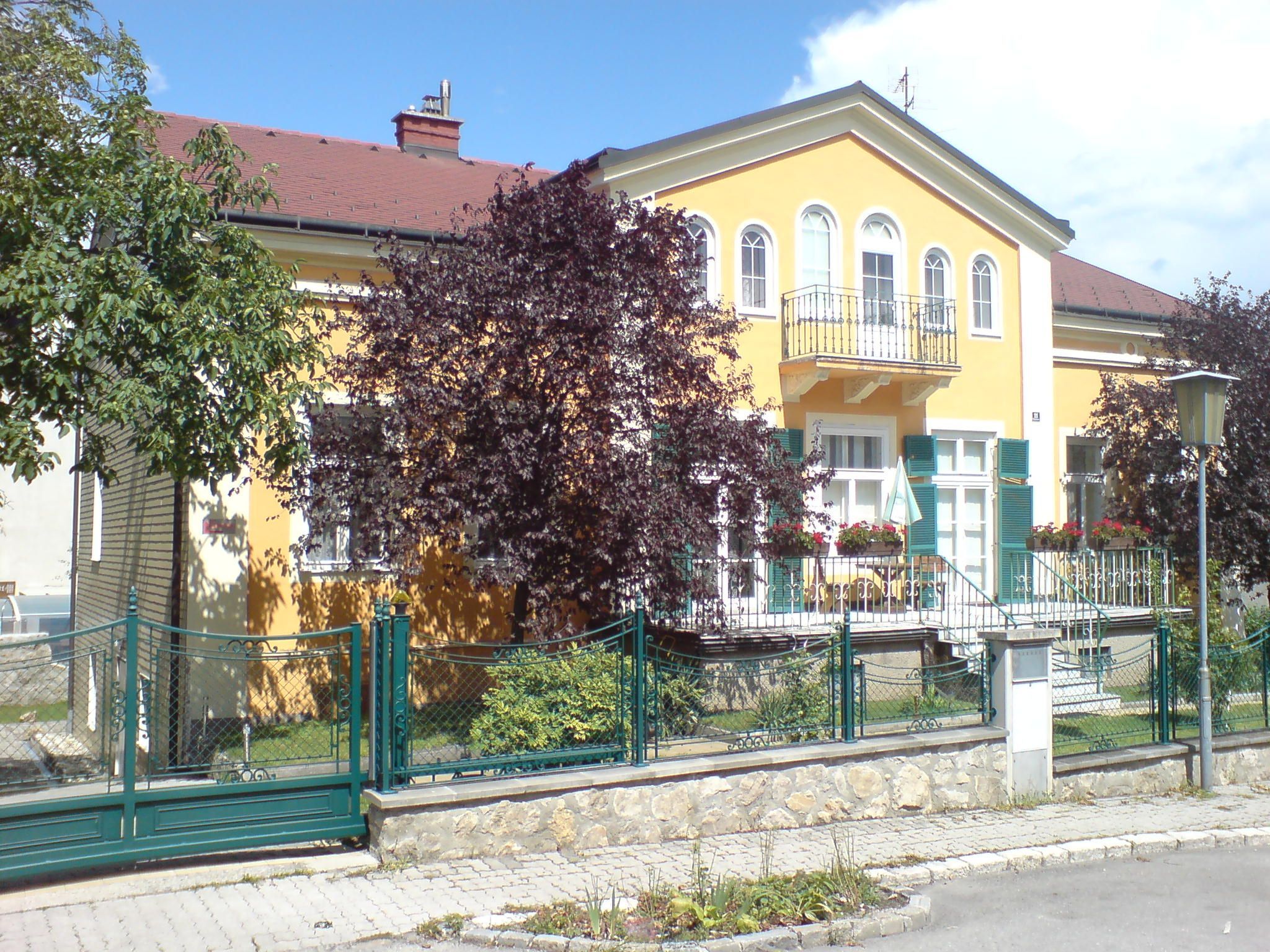 Žlutý dům s balkonem a zeleným plotem, obklopený stromy.