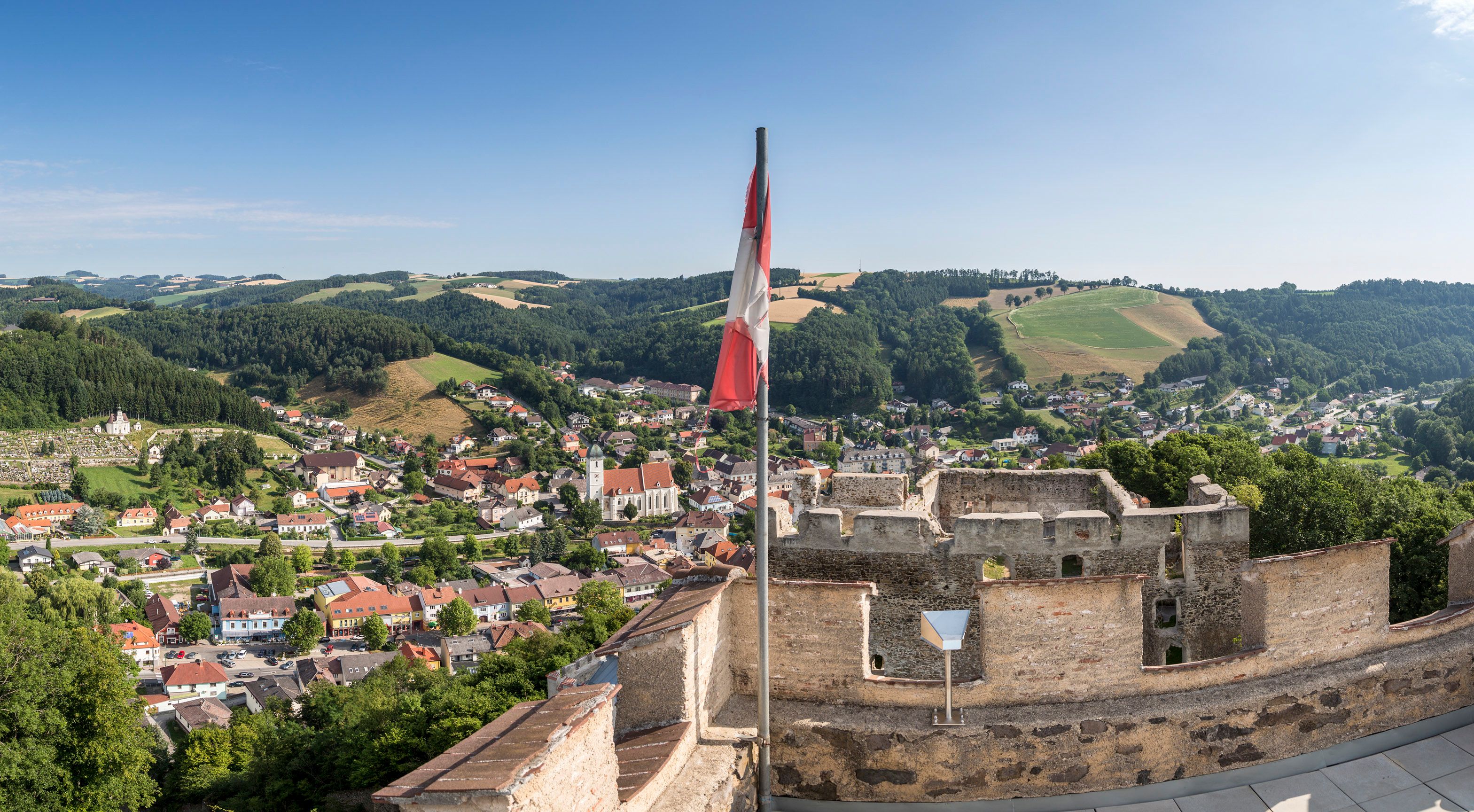 Panoramatický pohled ze zříceniny hradu na město s kopci v pozadí a rakouskou vlajkou v popředí.