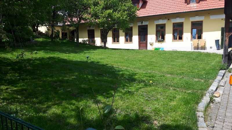 Zelená zahrada před žlutou budovou s červenými střešními taškami a několika okny.