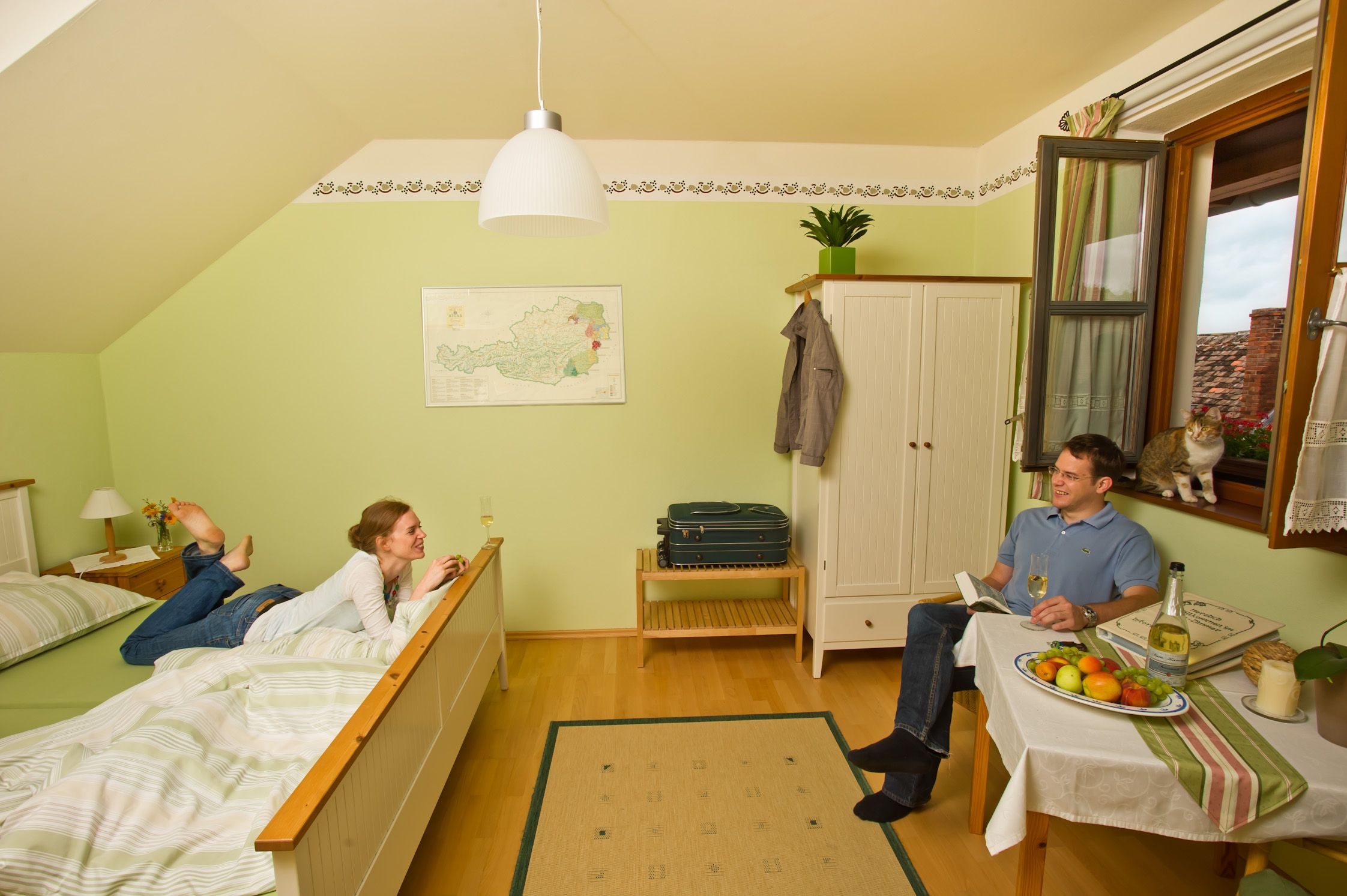 Útulný pokoj pro hosty s relaxující dvojicí. Žena leží na posteli, muž sedí u stolu. U okna sedí kočka.