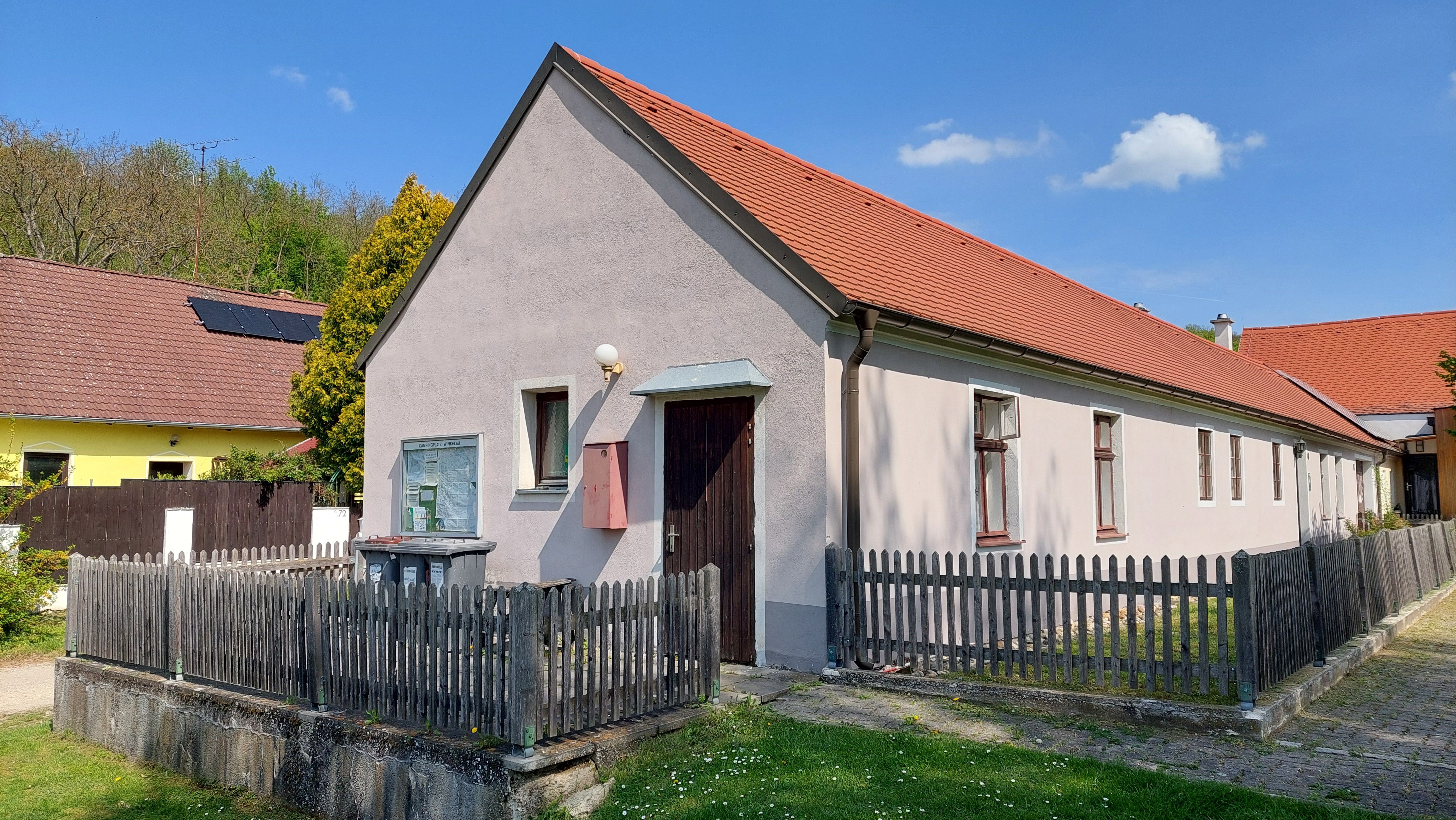 Dlouhý jednopatrový dům s červenou střechou a dřevěným plotem, obklopený zelenou trávou a modrou oblohou.