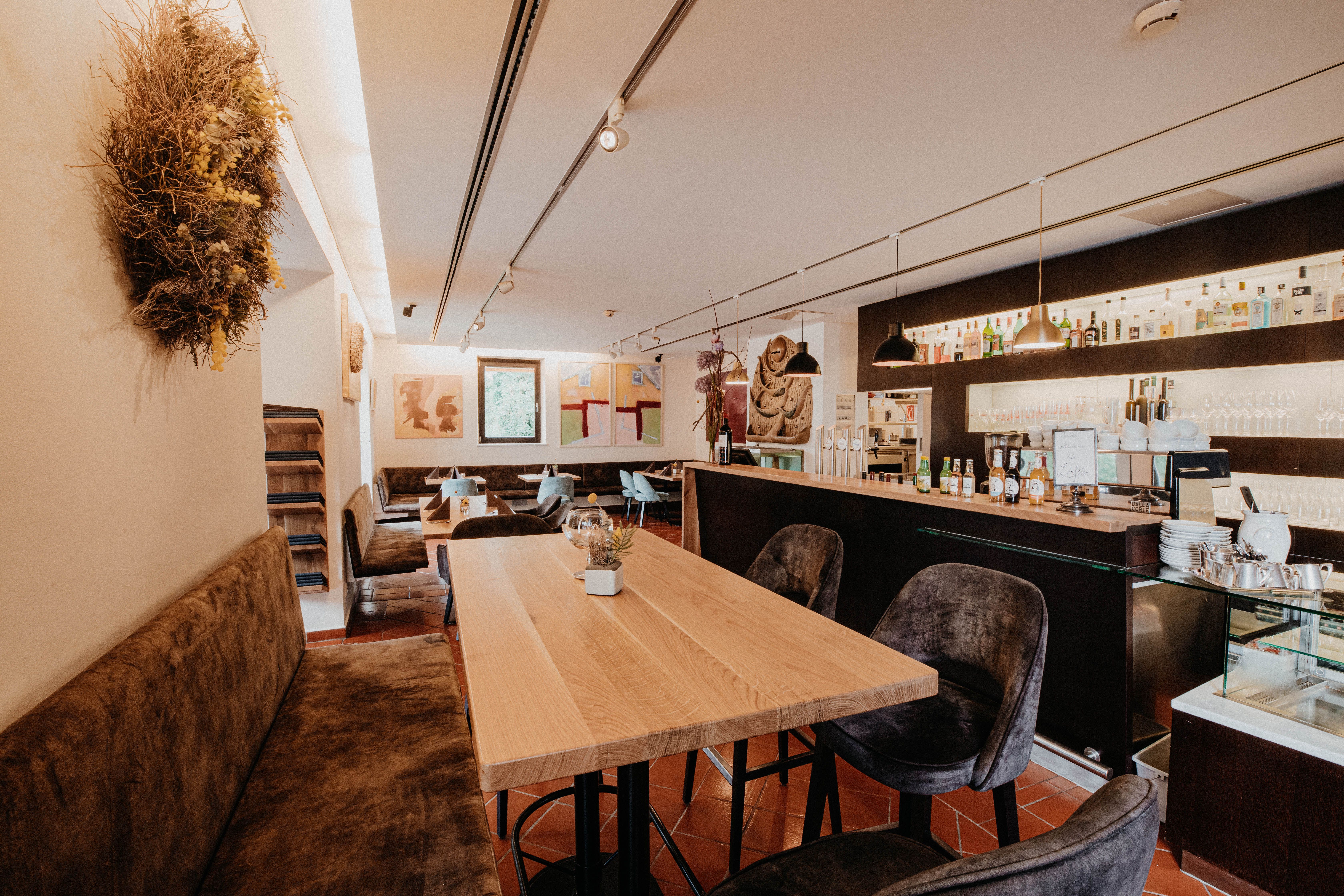 Pohled do interiéru moderní restaurace s dřevěnými stoly, barem a uměleckými díly na stěnách.