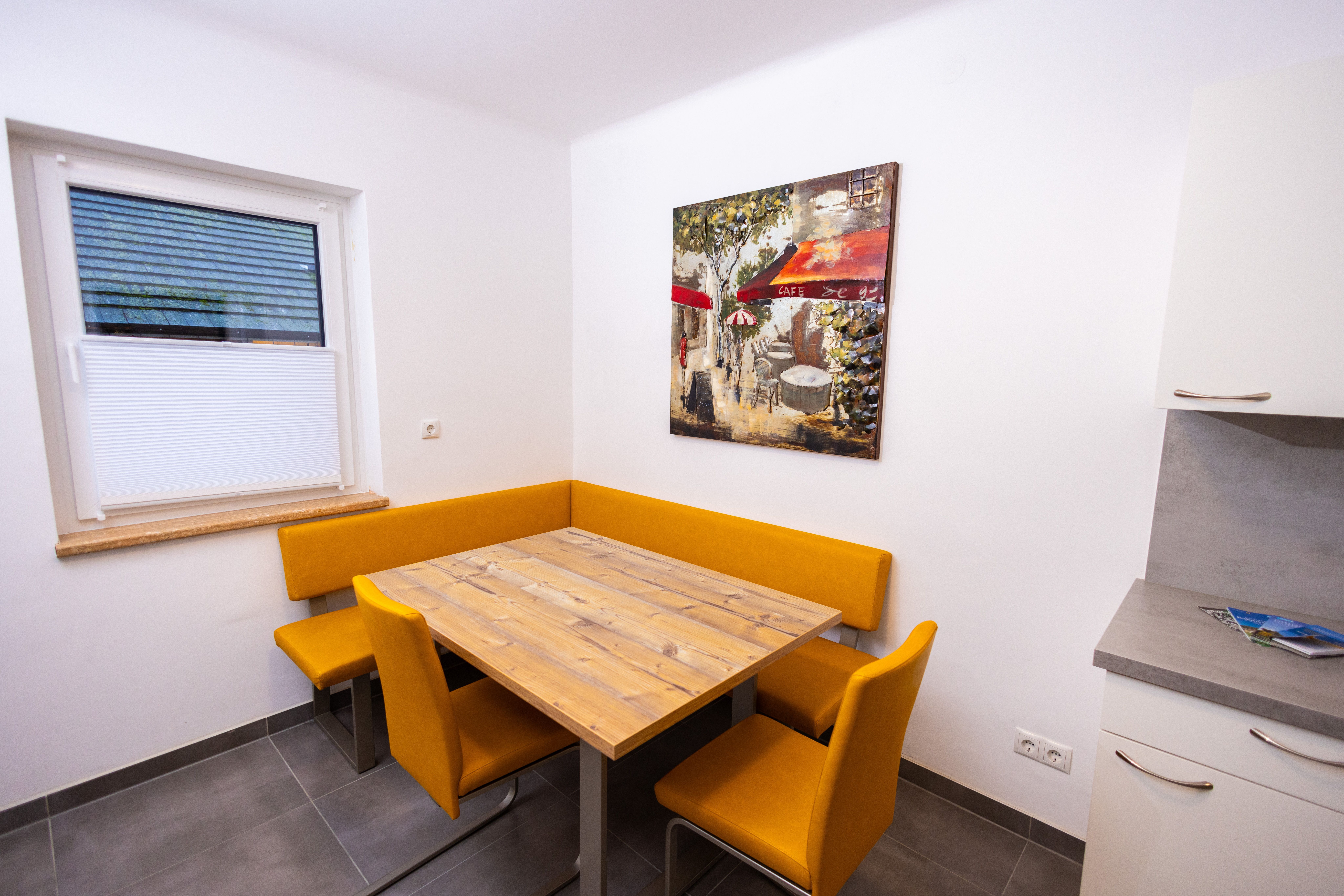 Jídelní kout s dřevěným stolem, žlutými židlemi a nástěnnou malbou kavárny.