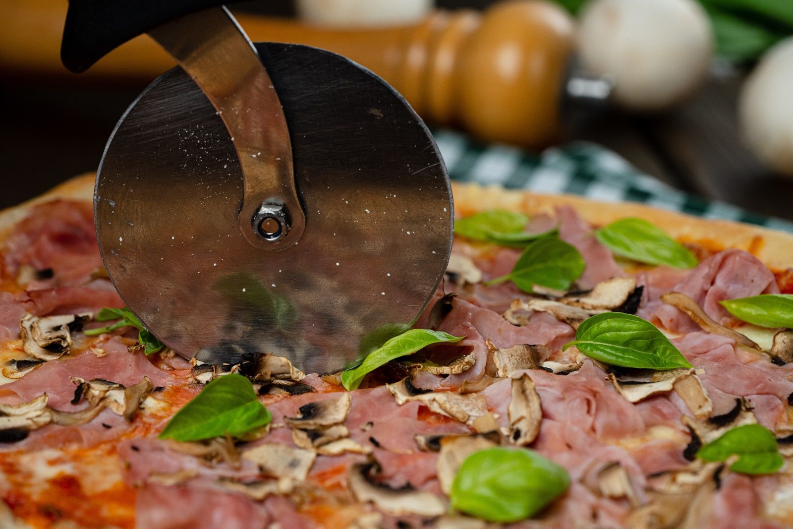 Detailní záběr pizzy se šunkou, žampiony a bazalkou, která se krájí nožem na pizzu.