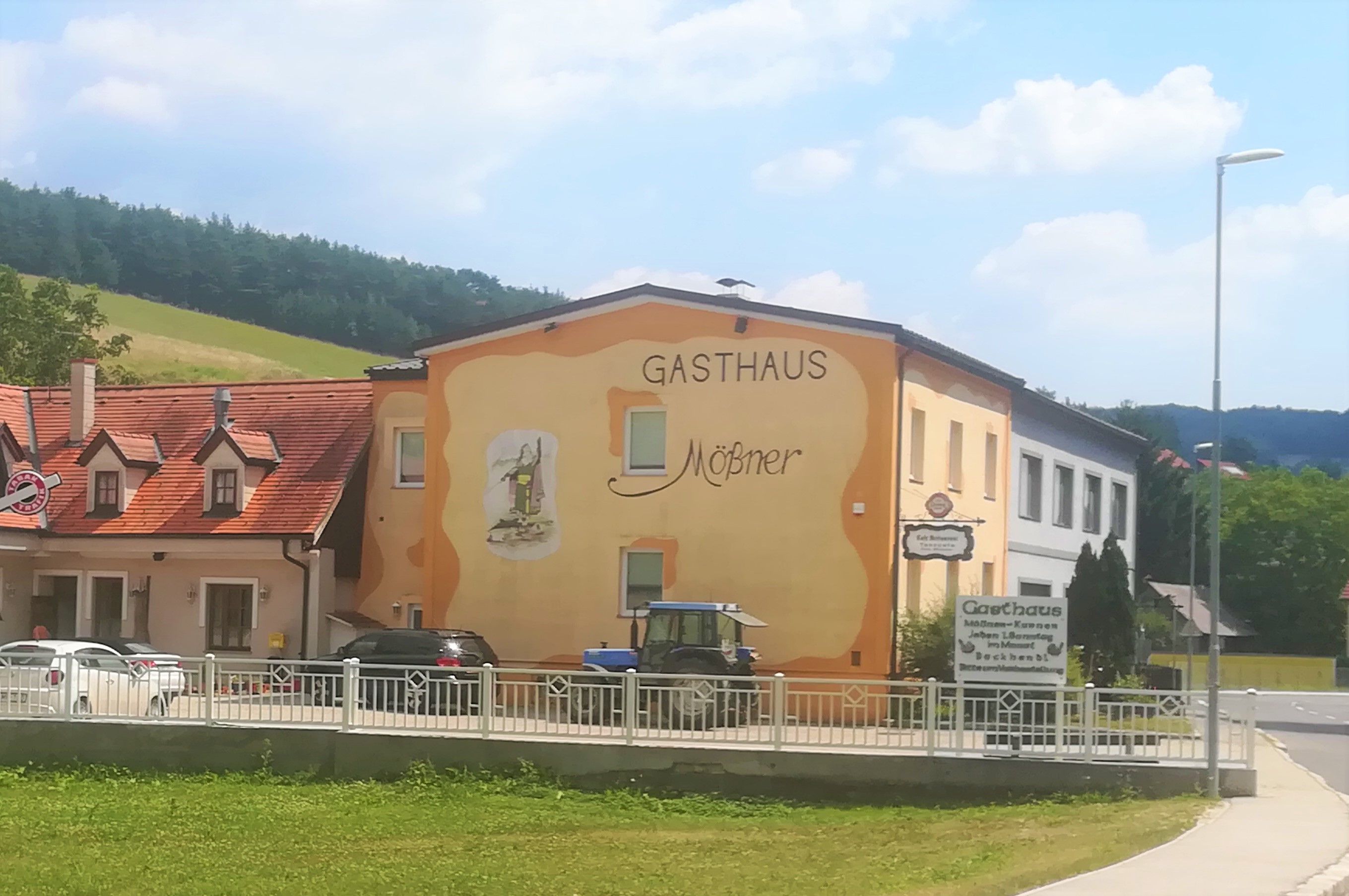 Exteriérový pohled na žlutooranžově natřený hostinec Mößner ve venkovském prostředí s modrým traktorem a auty před budovou.