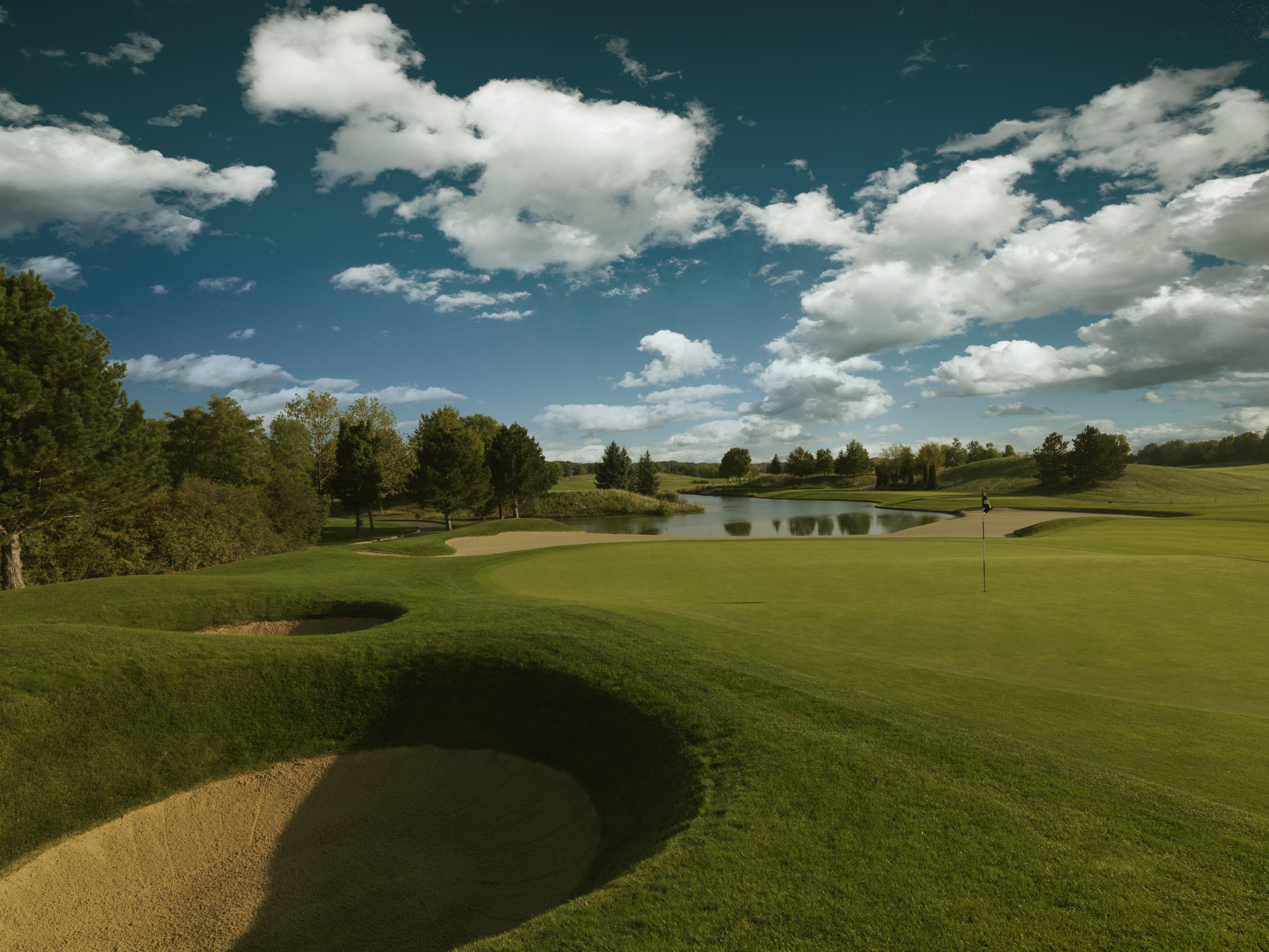 Golfové hřiště s písečnými bunkry, greenem a rybníkem pod modrou oblohou s mraky.