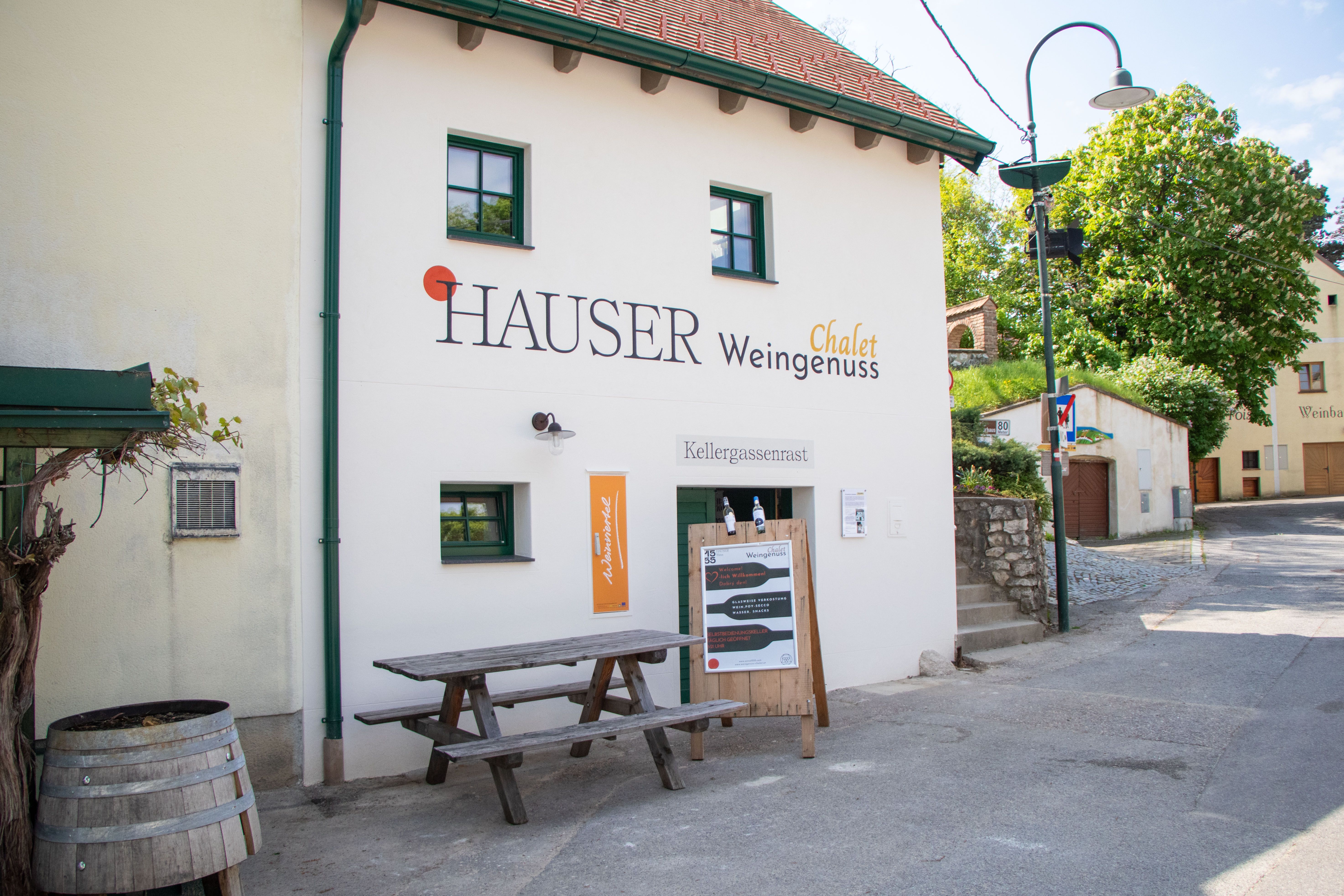 Pohled zvenčí na budovu s označením "Hauser Chalet Weingenuss". Před budovou stojí dřevěný stůl a sud na víno.