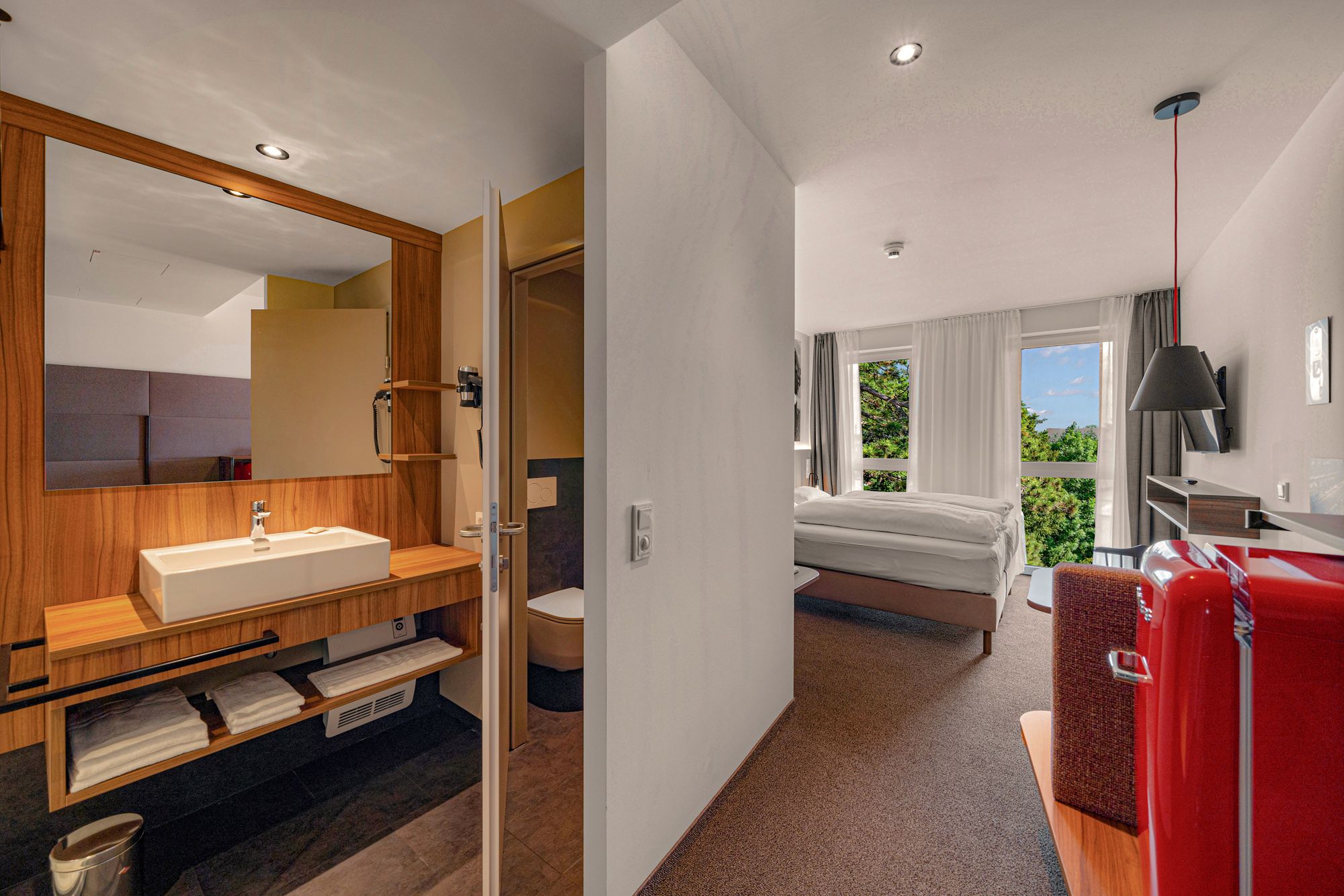 Pohled do interiéru moderního hotelového pokoje s koupelnou, postelí a červenou lednicí.