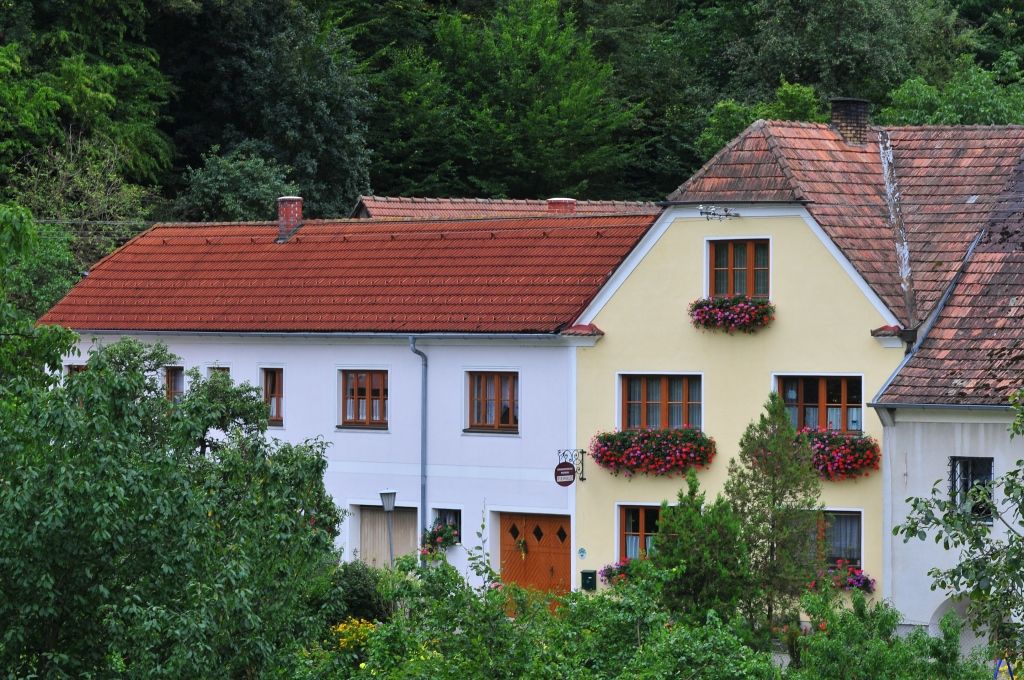 Tradiční dům s červenými střešními taškami a květinovými truhlíky před okny, obklopený zelení.
