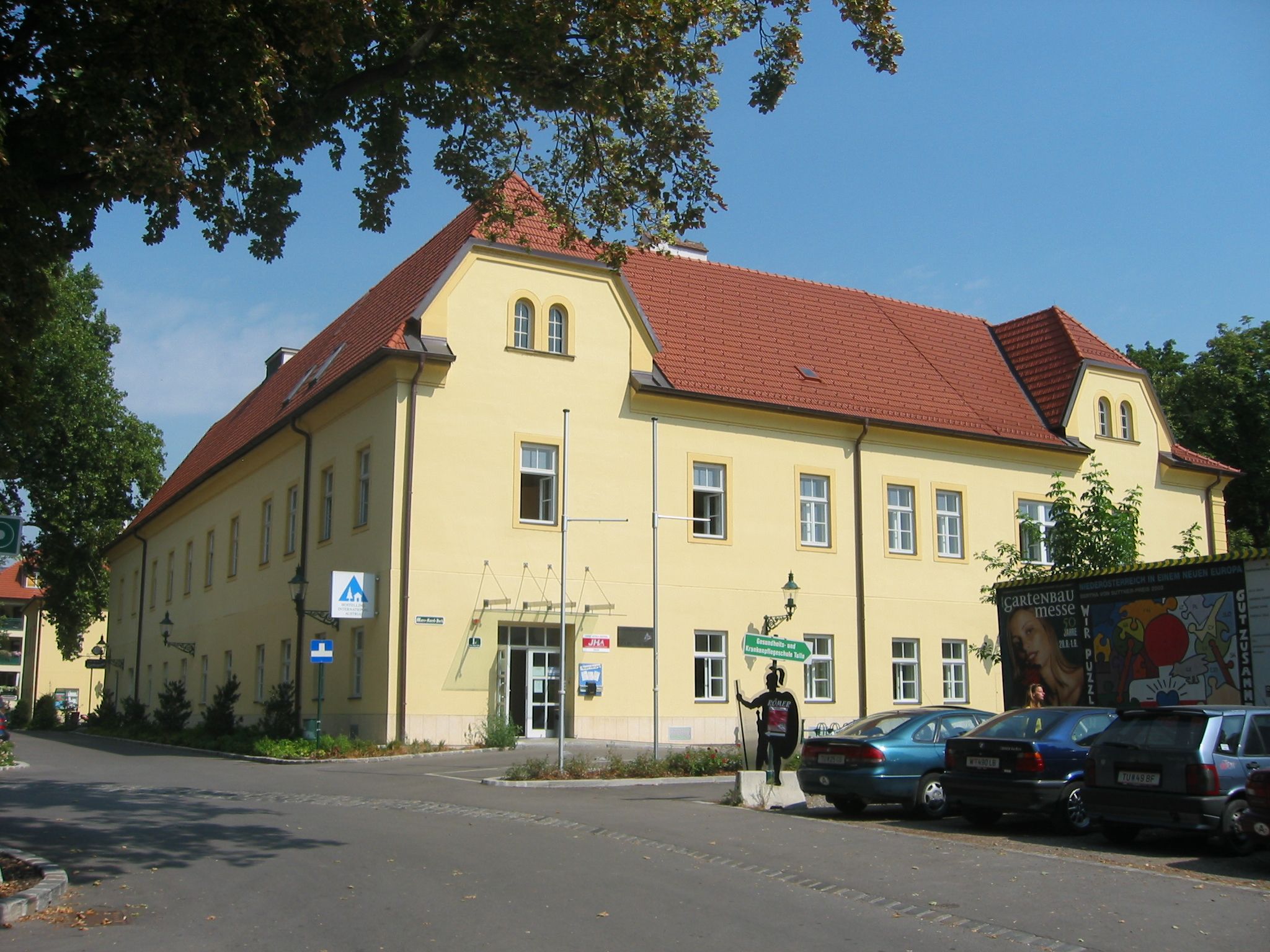 Pohled zvenčí na žlutou budovu s červenou střechou, Junges Hotel Tulln, obklopenou stromy a zaparkovanými auty.