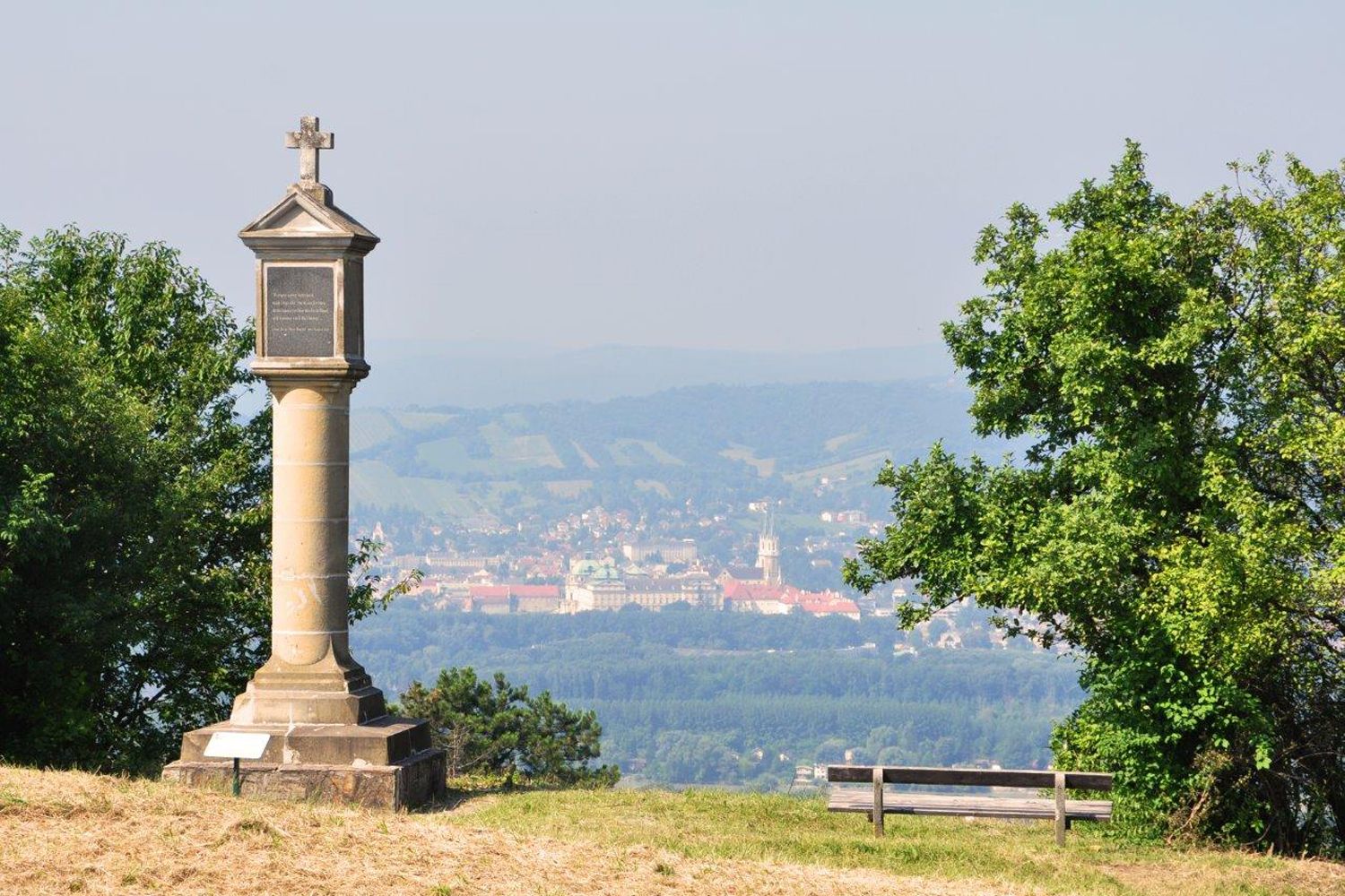 Sloup s křížem na kopci, v pozadí město a kopce.