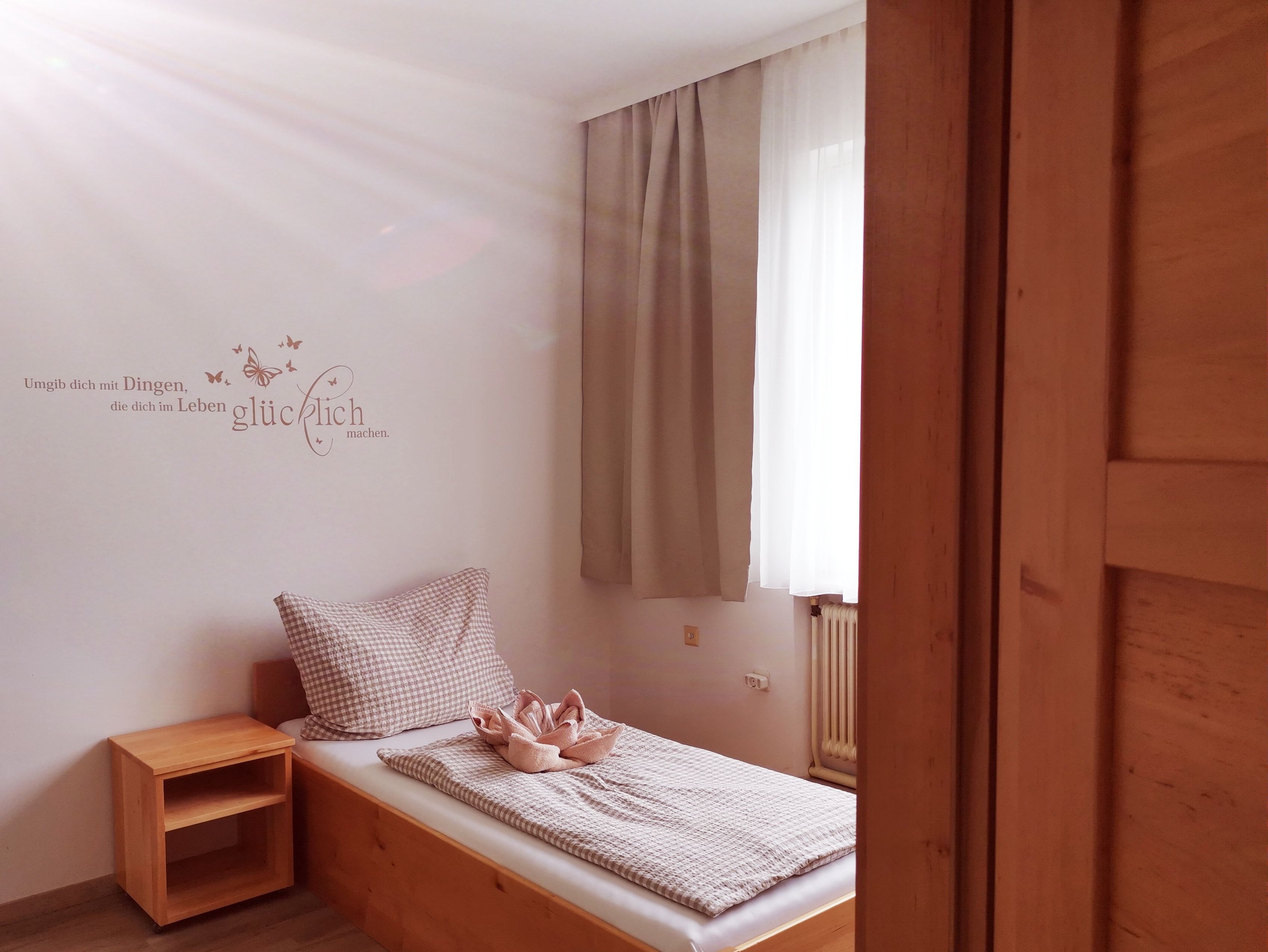 Jednoduchý pokoj pro hosty s jednou postelí, dřevěnými policemi a citátem na zdi.