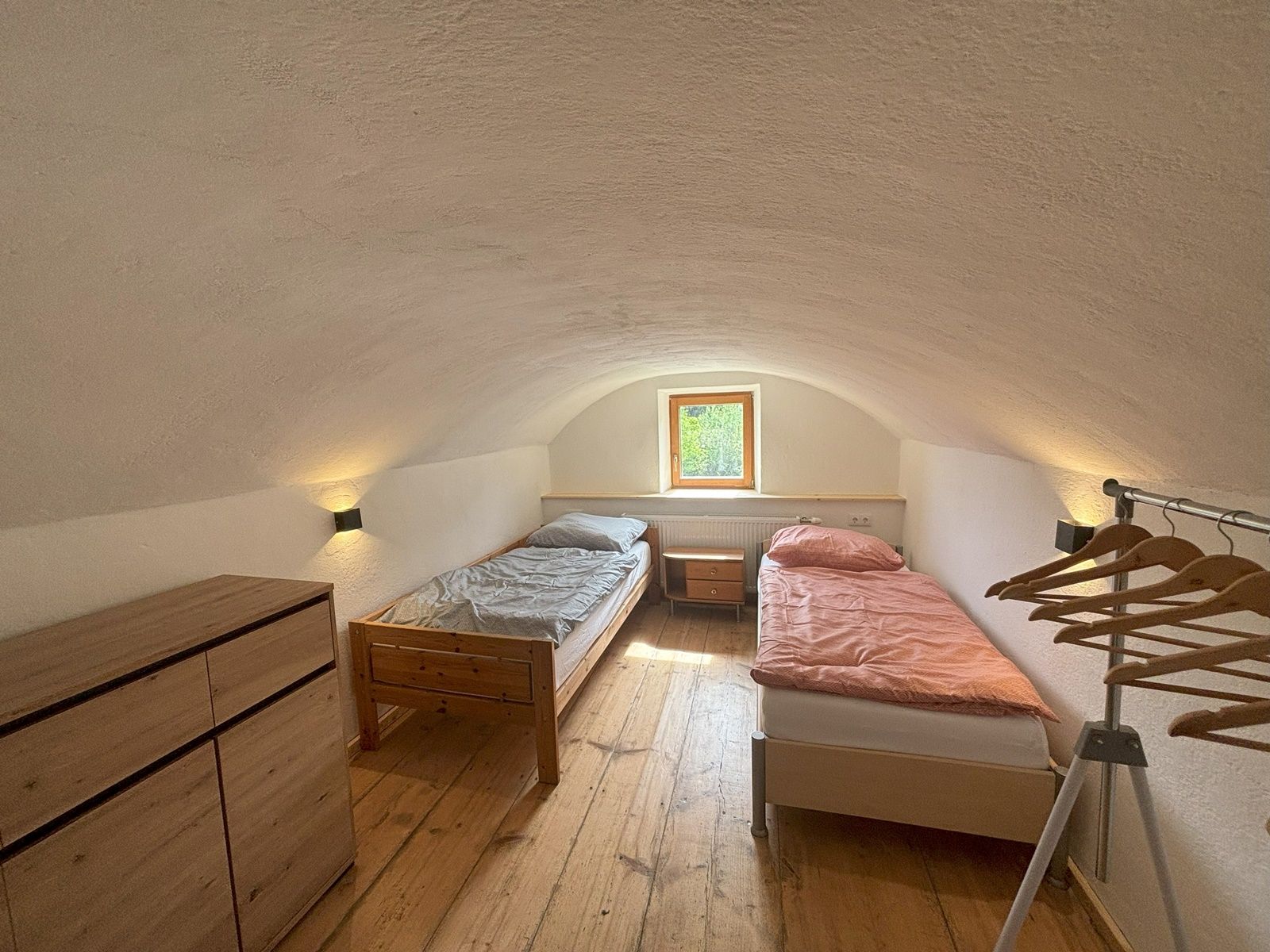 Jednoduchý dvoulůžkový pokoj se dvěma samostatnými lůžky, komodou a věšákem na oblečení. Malé okno propouští přirozené světlo.