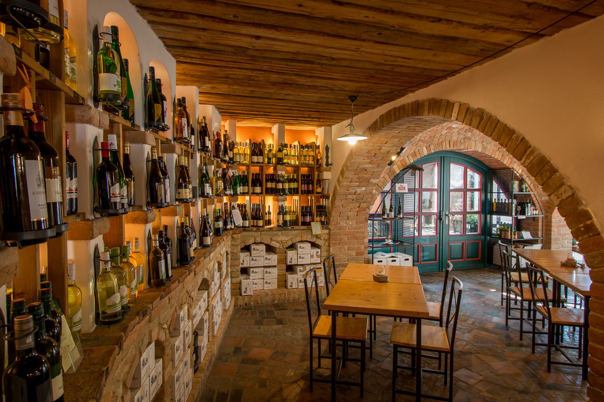 Vinný sklípek s policemi plnými lahví vína, dřevěnými stoly a cihlovými oblouky.