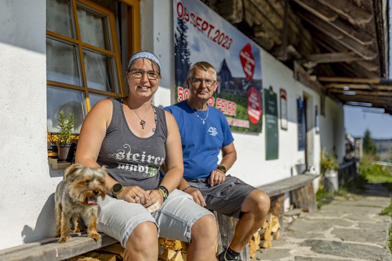 Dva lidé sedící před budovou se psem, v pozadí plakát s datem 09. září 2023.