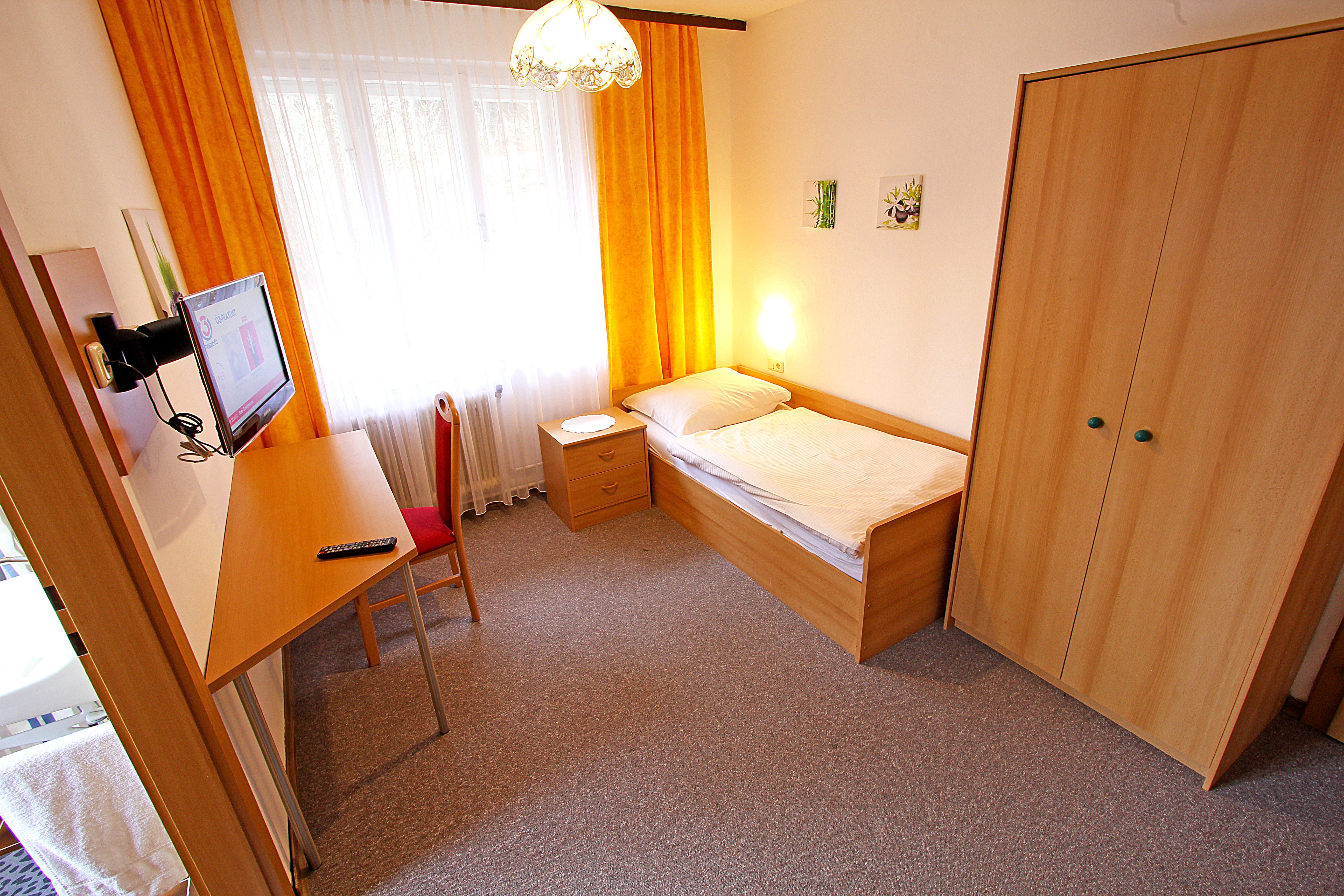 Jednoduše zařízený jednolůžkový pokoj s postelí, psacím stolem, židlí, skříní a TV. Žluté závěsy a koberec na podlaze.