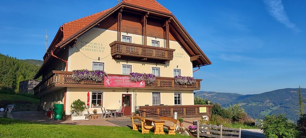 Tradiční hostinec s dřevěnými balkony a květinami, obklopený zelenými kopci a modrou oblohou.